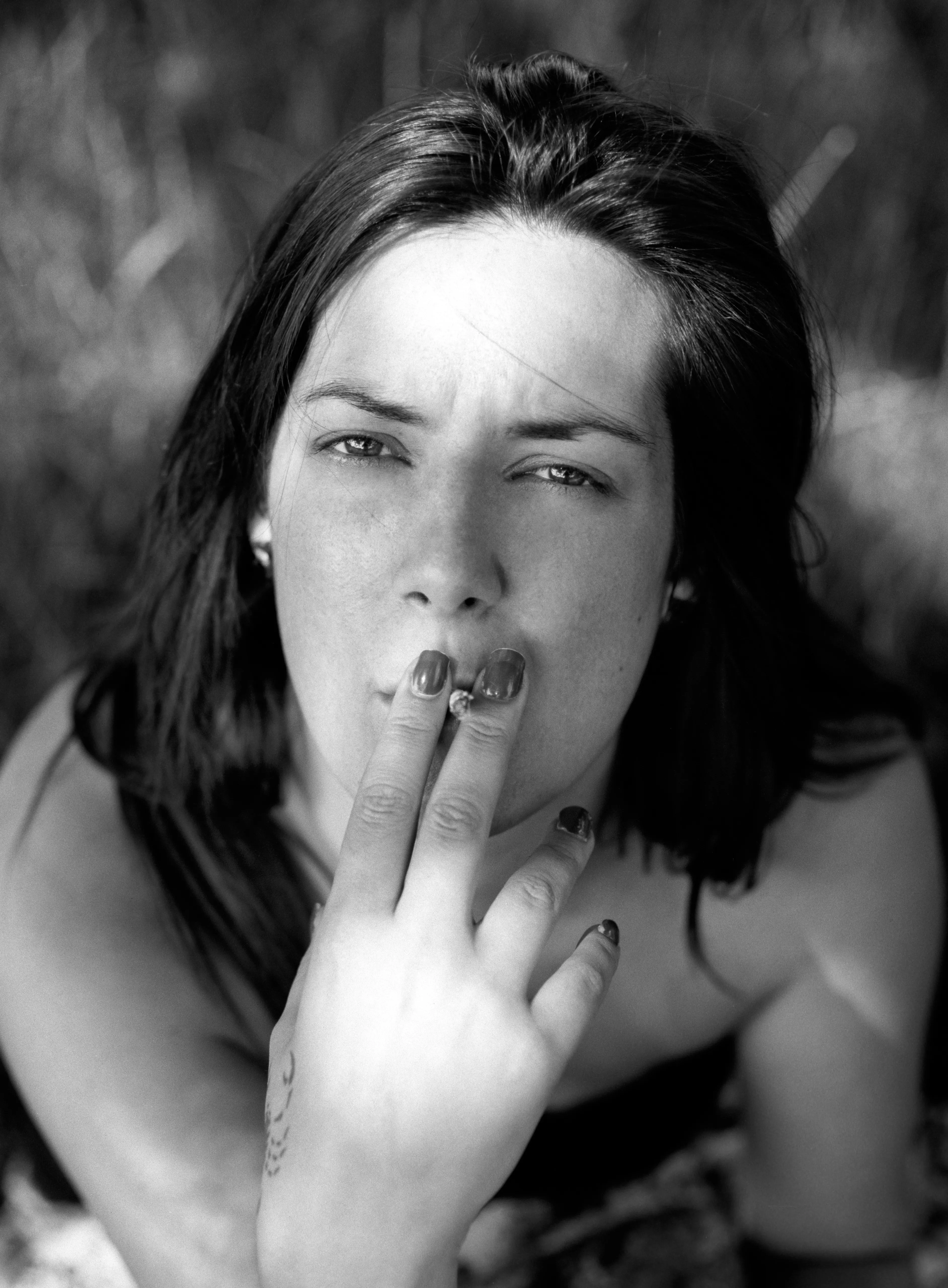 Black-and-white portrait of a woman in long grass drawing on a cigarette held to her lips, looking directly at the camera.