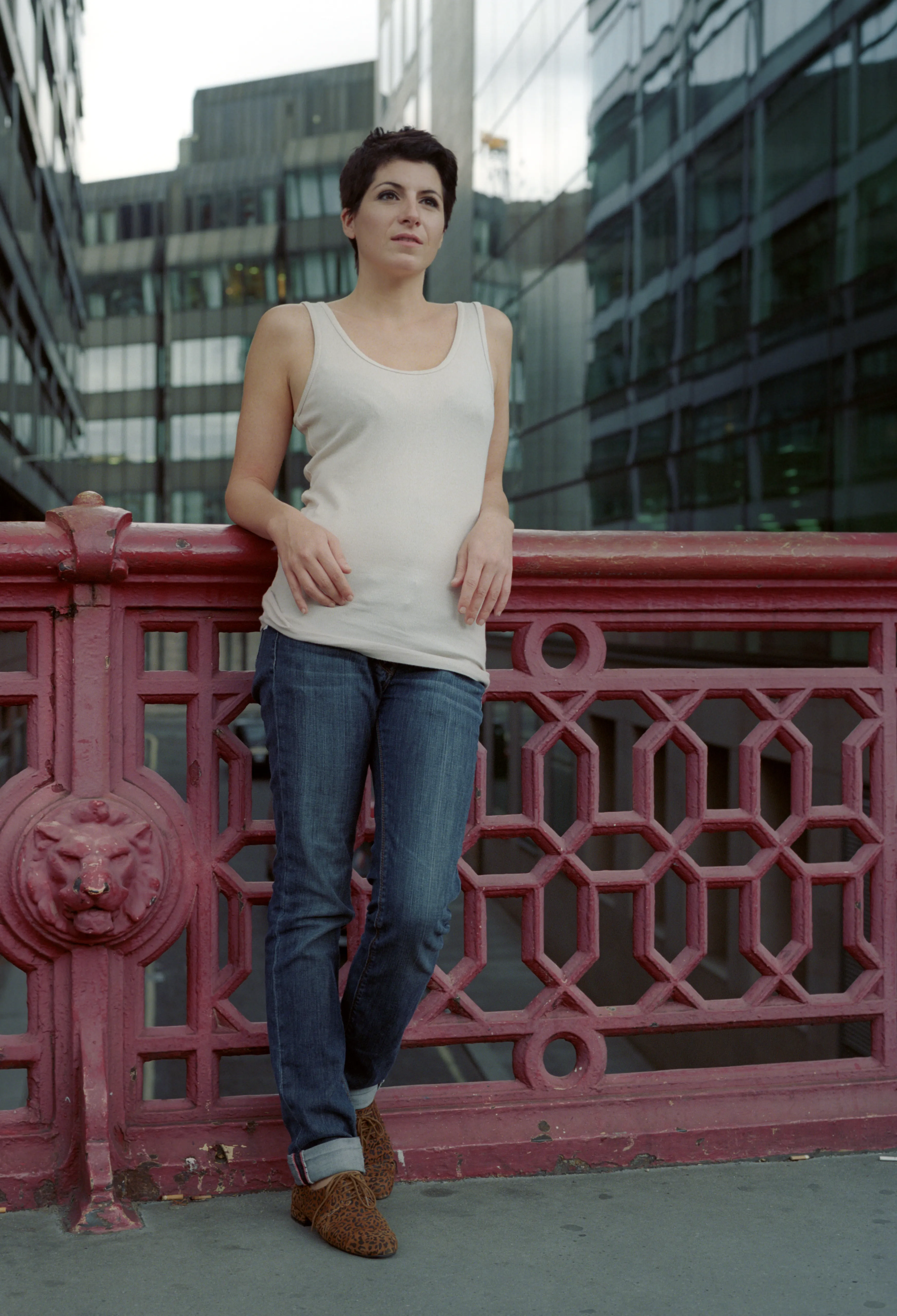 A woman in a white vest top and jeans leans against an ornate red-painted iron bridge railing, looking to one side; office buildings rise behind her.