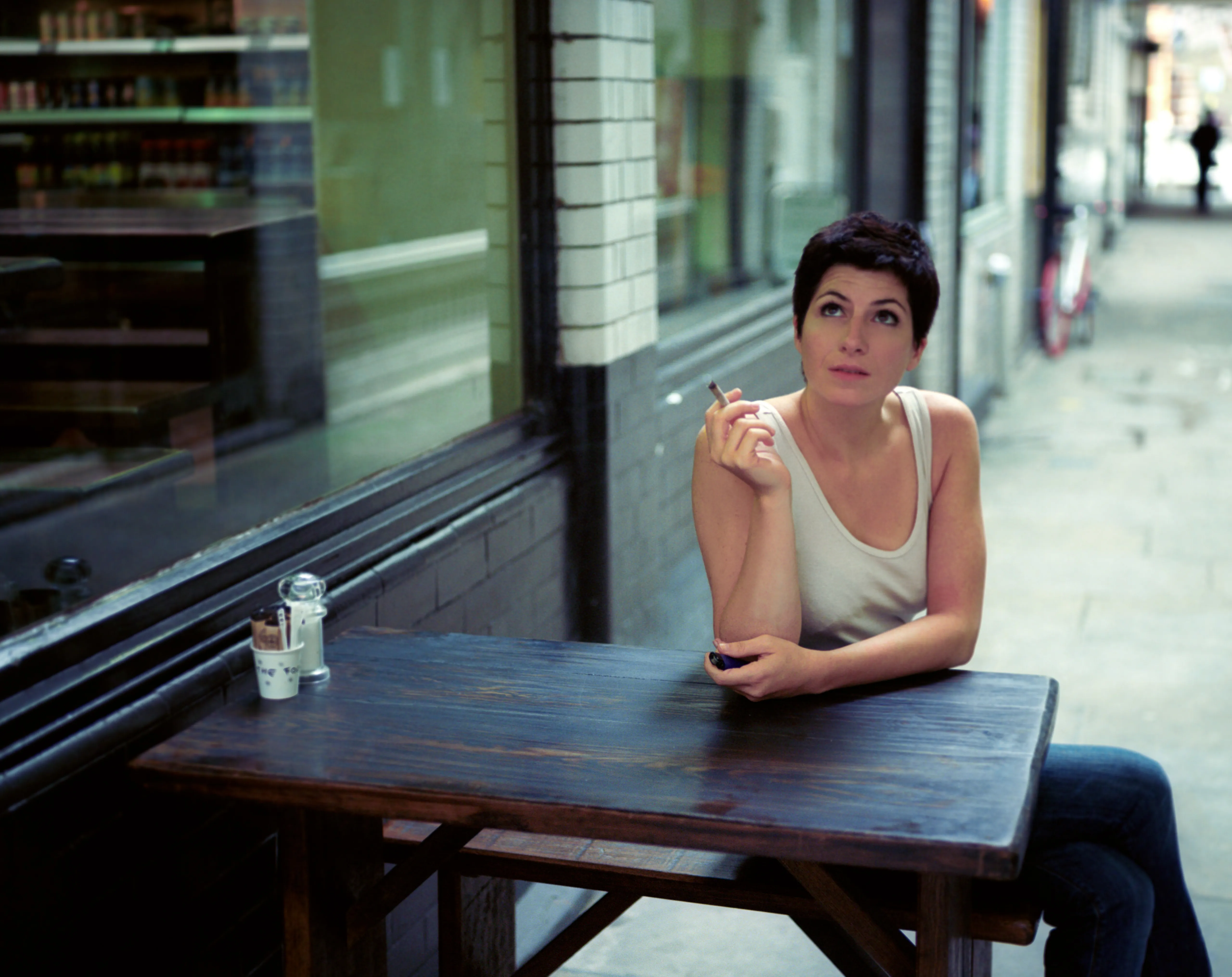 A woman sits alone at a wooden table outside a shopfront, holding a cigarette and looking upward; a lighter rests on the table.