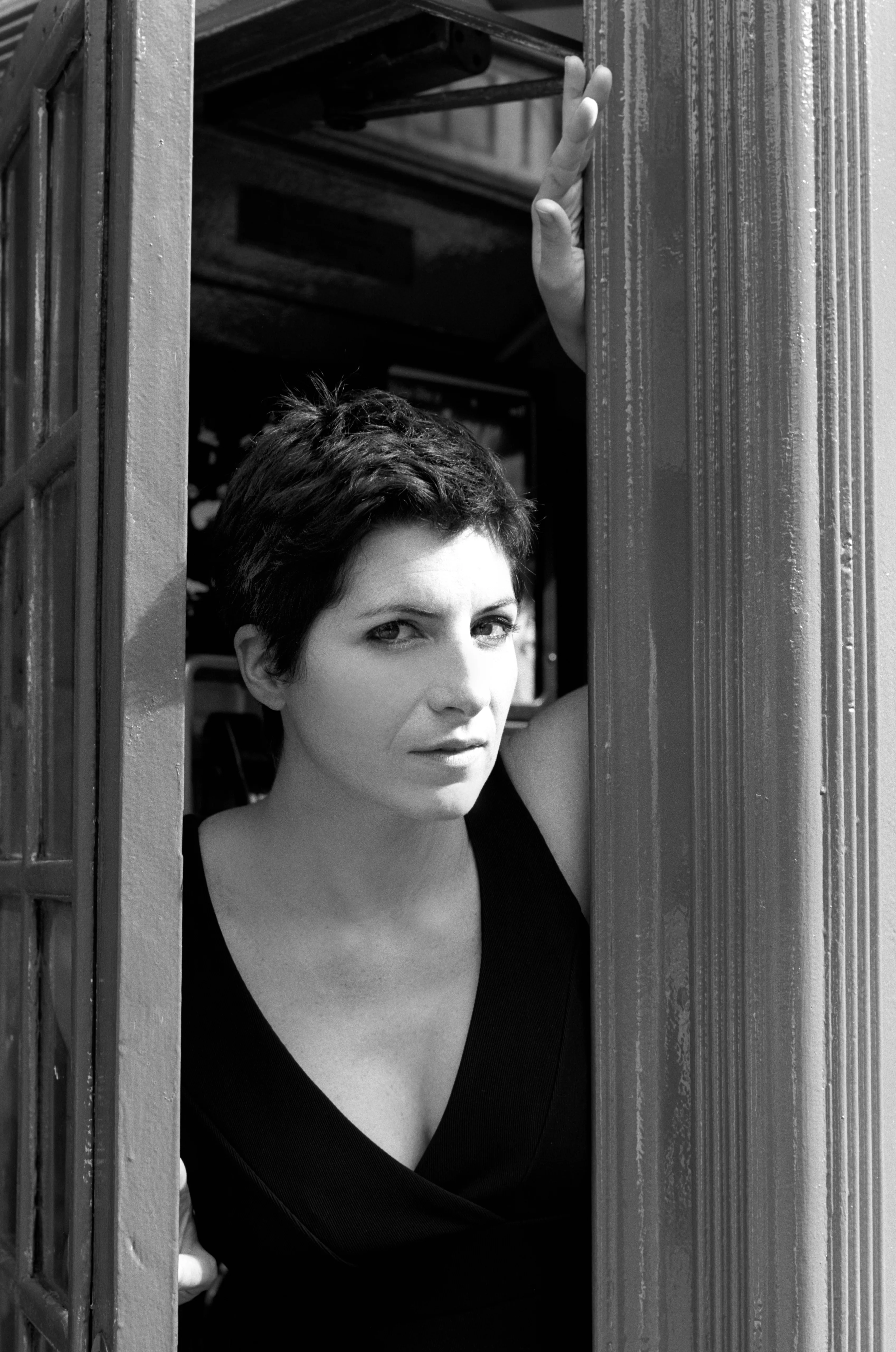 A woman with short dark hair in a black sleeveless dress stands in an open doorway, one hand raised against the painted frame, looking out towards the camera.