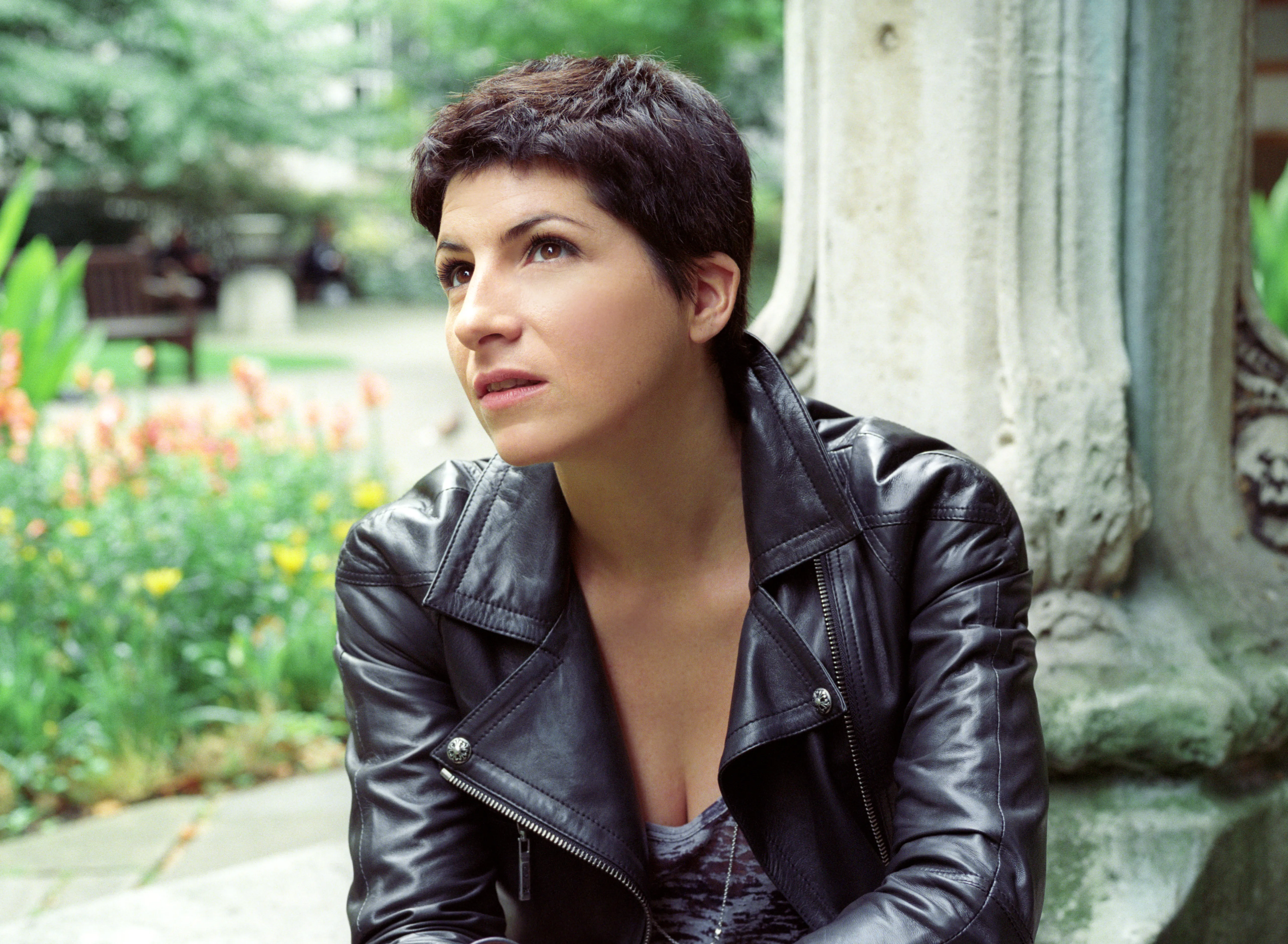 A woman in a black leather jacket sits beside a stone plinth, looking upward, flowers and a garden behind her.
