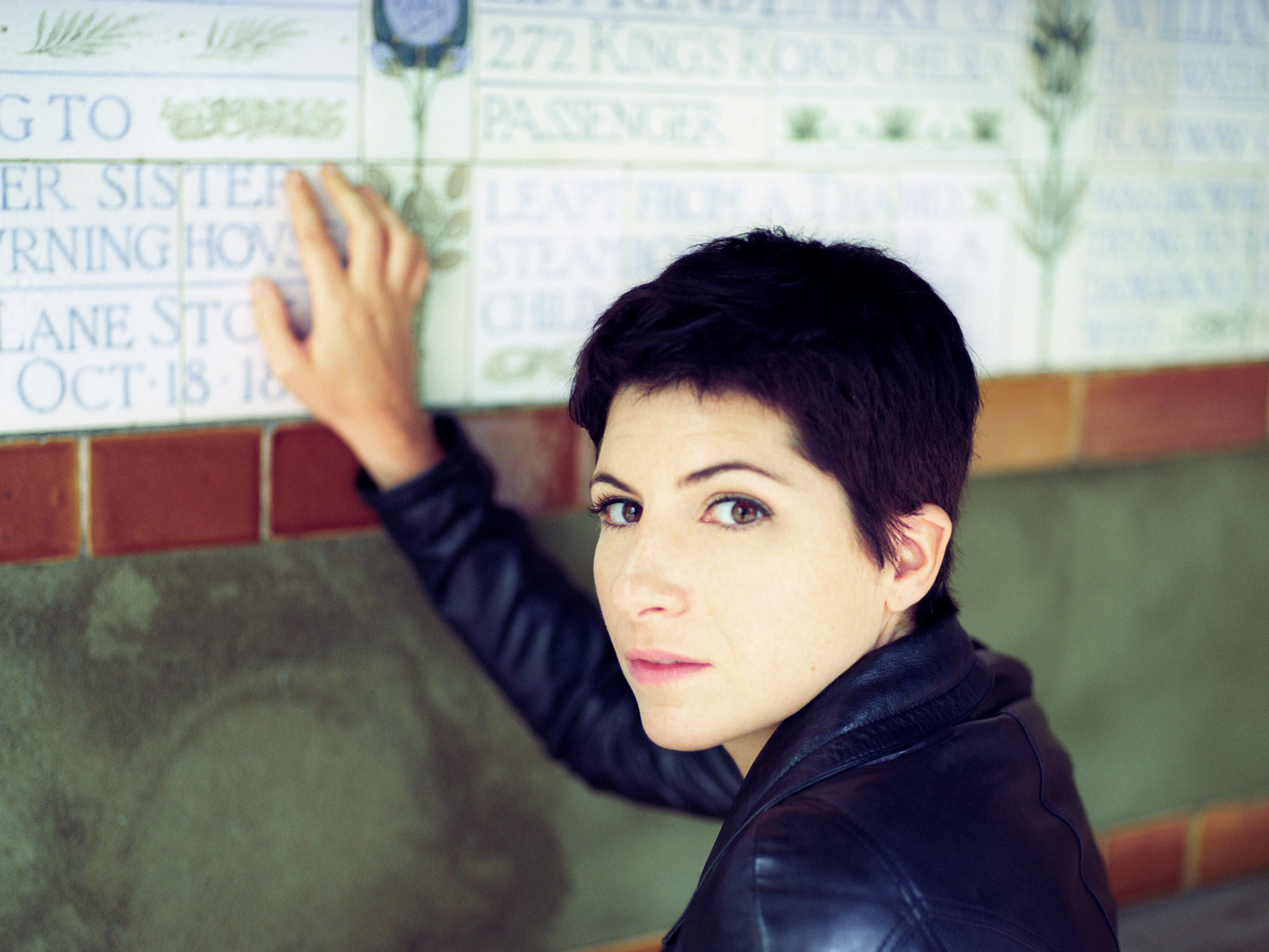 A woman reaches up to touch a lettered ceramic memorial tablet on a tiled wall, turning to look back over her shoulder towards the camera.