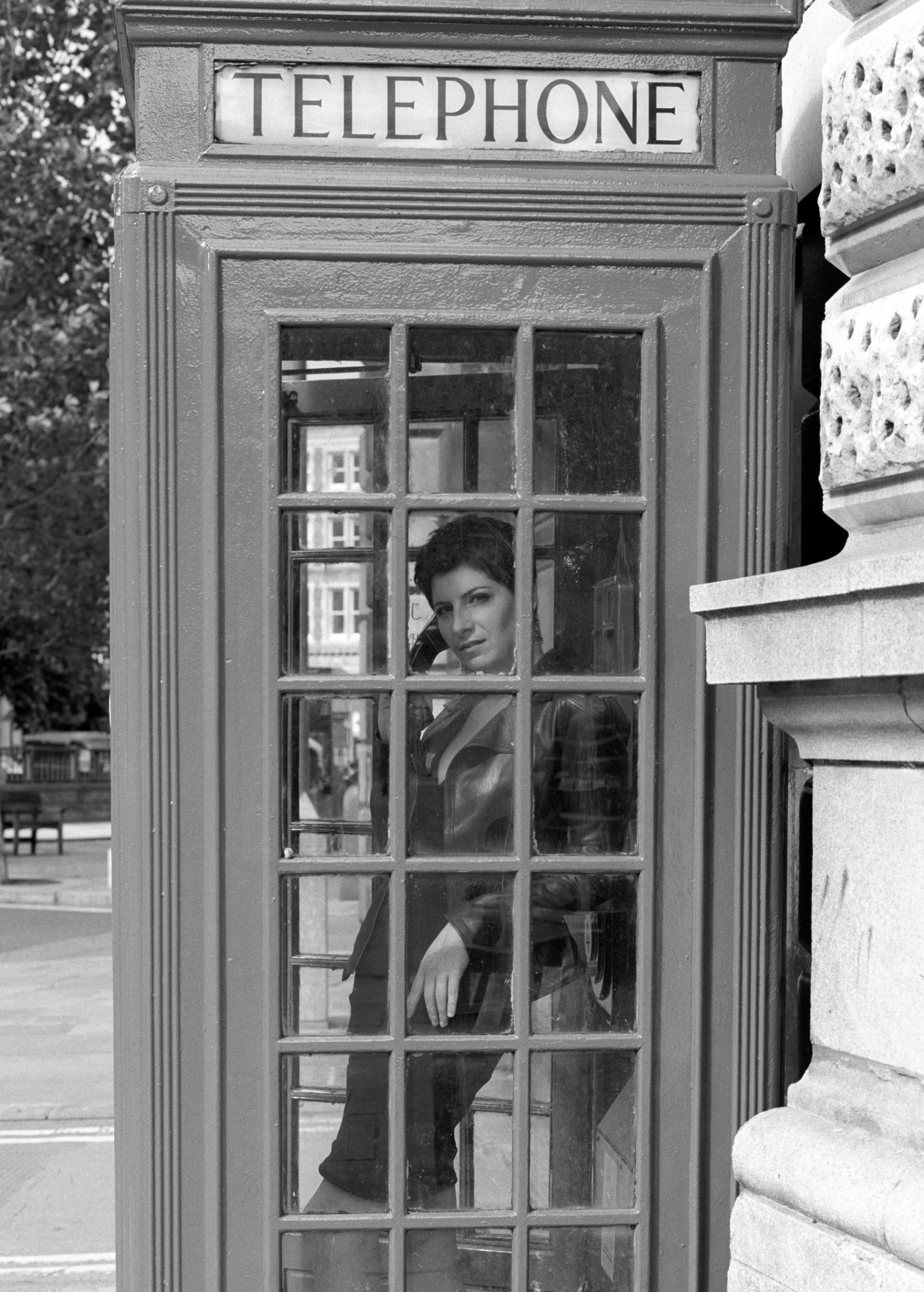 A woman stands inside a London telephone box, viewed through its glazed door, the word TELEPHONE lettered on the panel above.