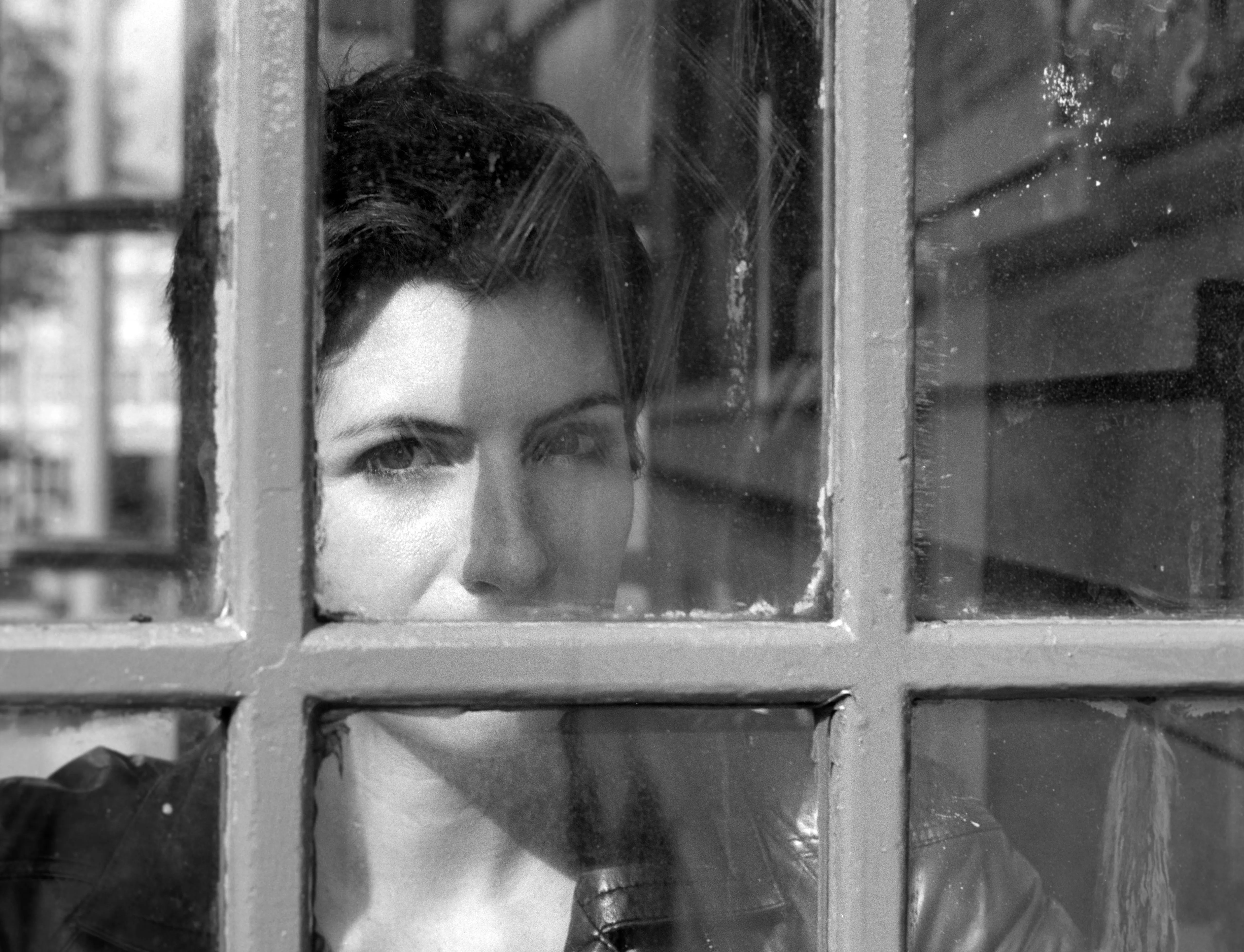 A woman's face seen through the scratched, grid-glazed window of a telephone box, looking downward, the street faintly reflected in the panes.