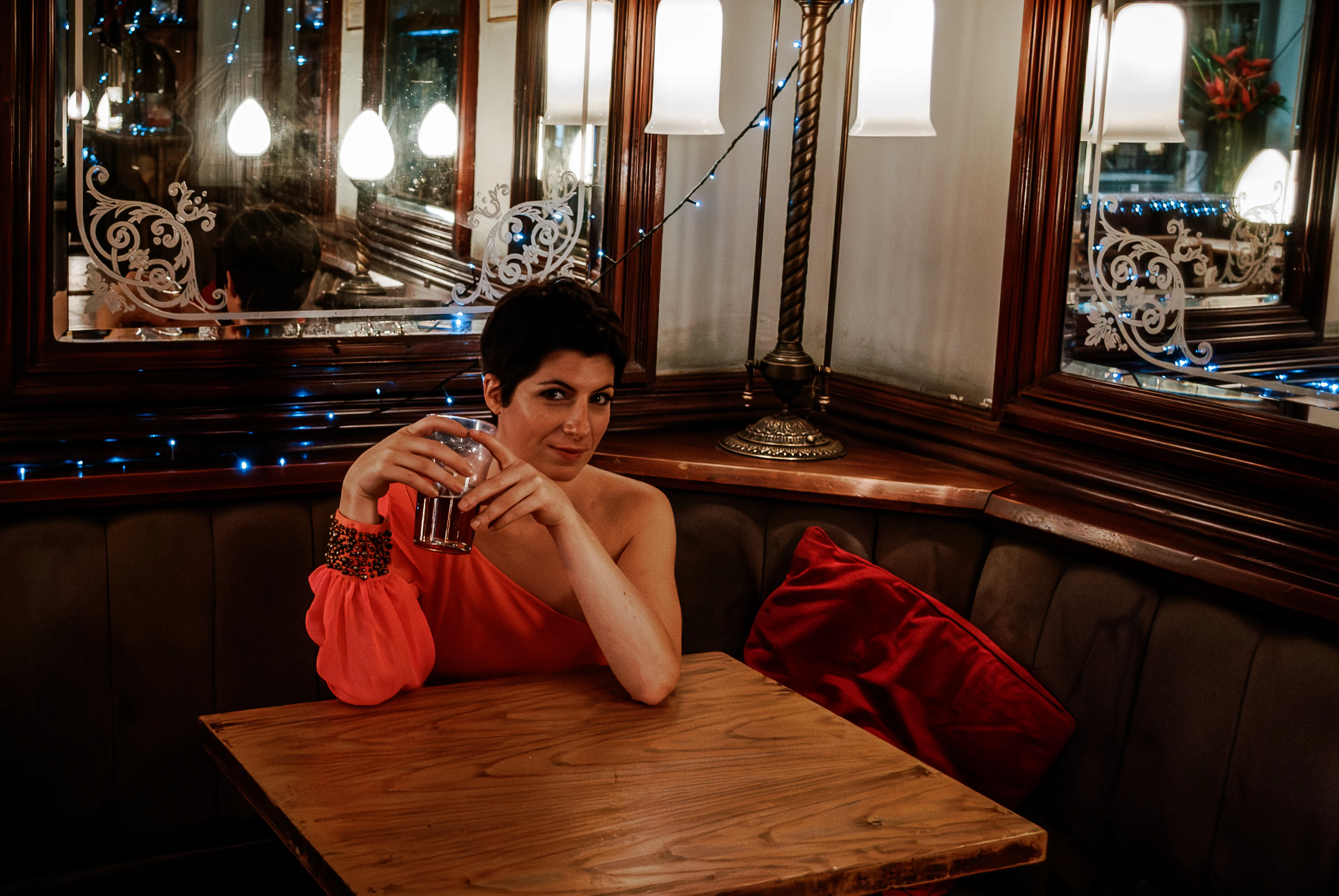 A woman in a coral one-shoulder top sits at a corner table in a pub, holding a glass with her chin resting on her hand; etched mirrors, fairy lights and a brass lamp surround her.
