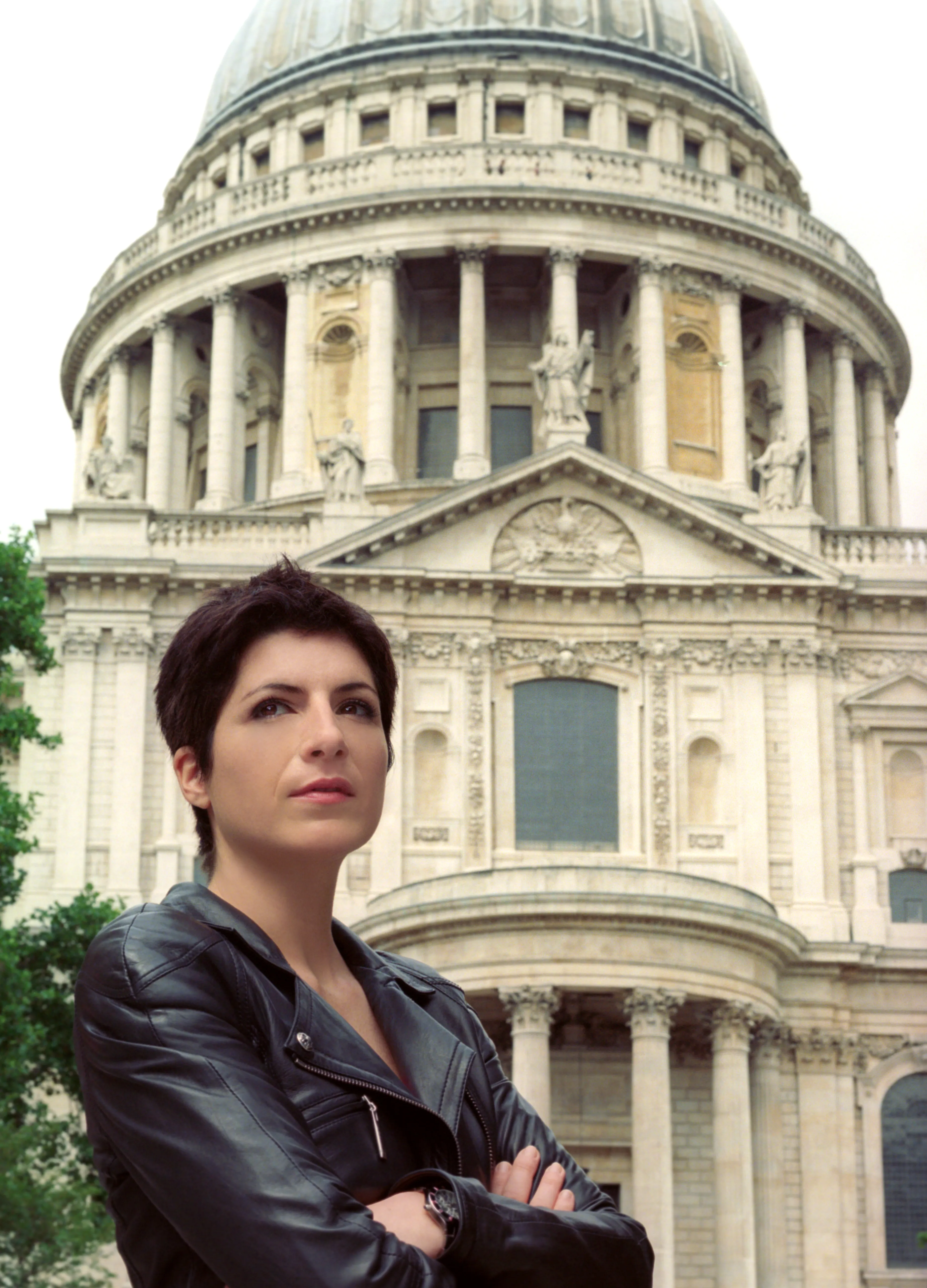 A woman in a black leather jacket stands with arms crossed below the dome and west front of St Paul's Cathedral, looking to one side.