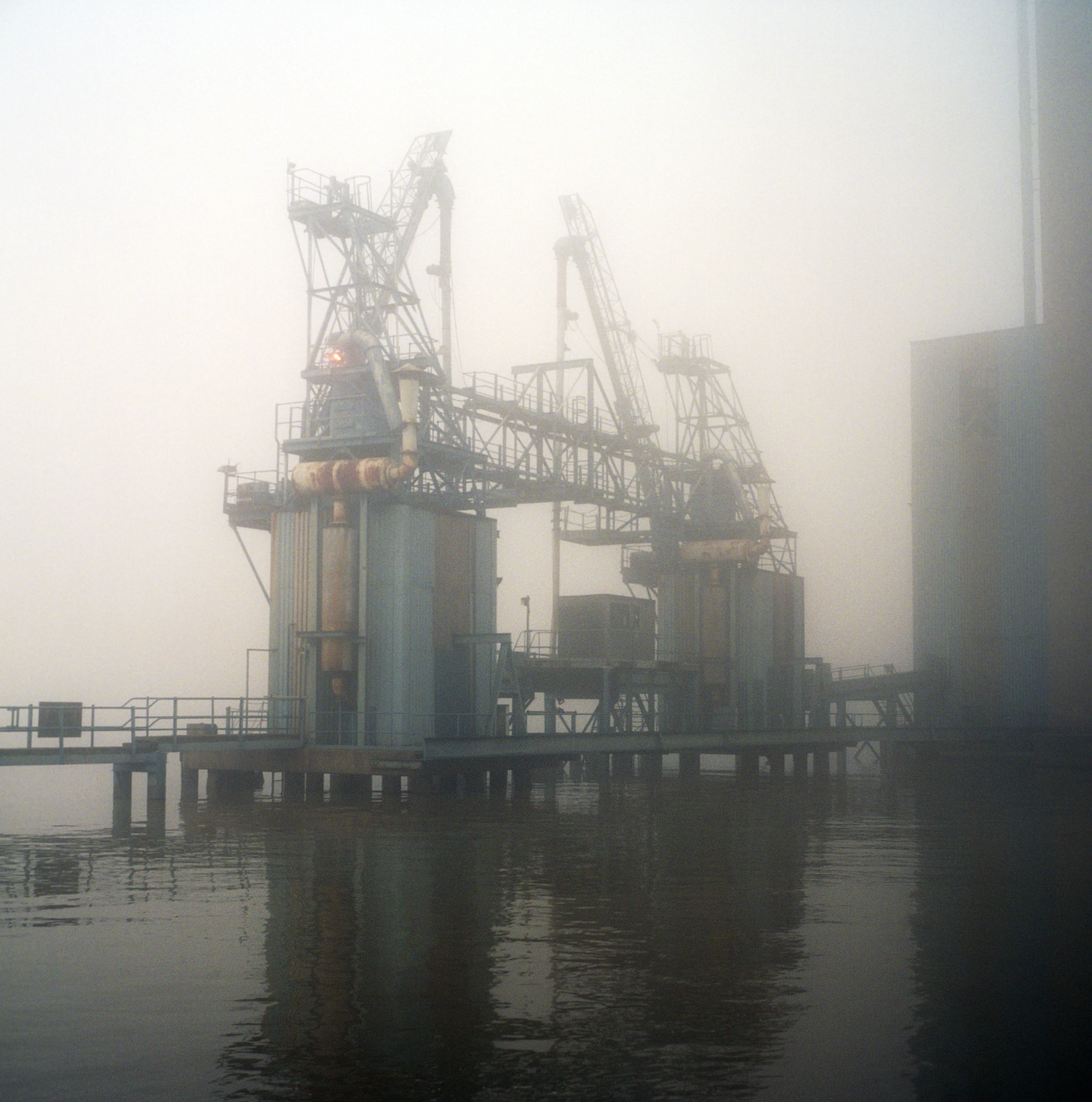Disused coaling cranes and loading gantries rising over still black water, dissolving into fog; a single red warning light glows on the structure.
