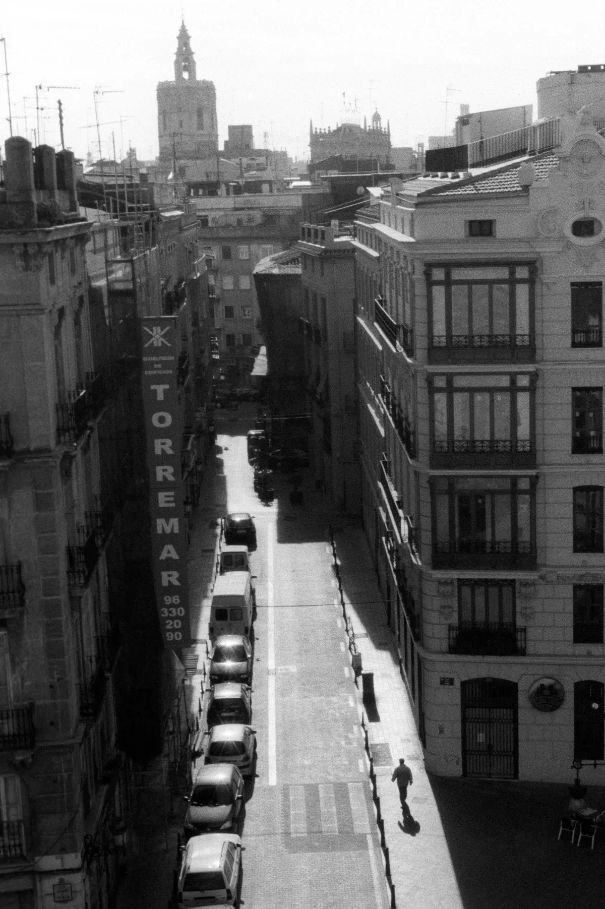 A narrow street from above, a man walking alone in the morning sun; the Miguelete on the skyline.