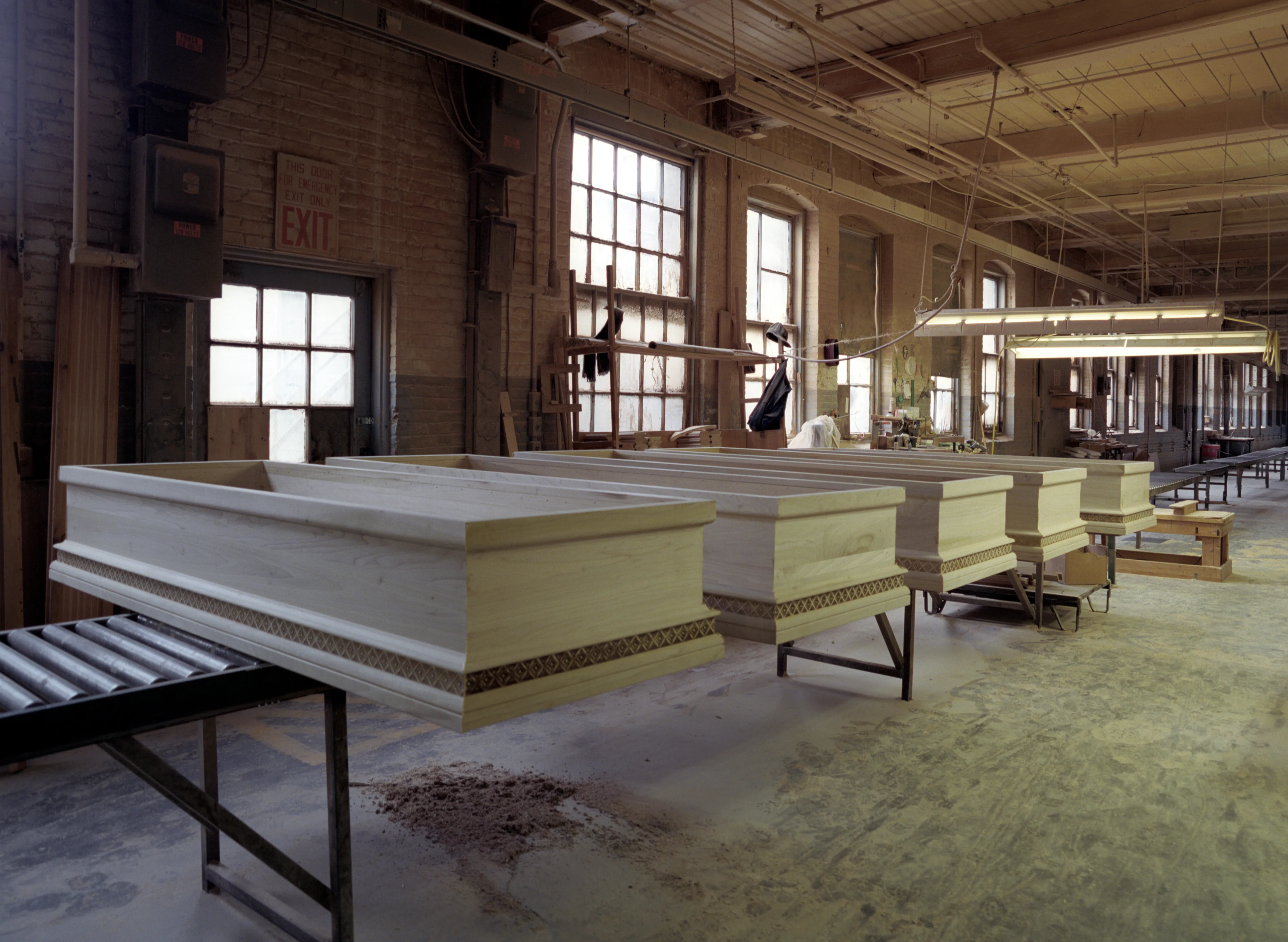 Five lidless casket shells in unstained wood recede along low trestles, each banded with embossed moulding; a roller conveyor enters at lower left, the tall windows behind blown white with daylight.