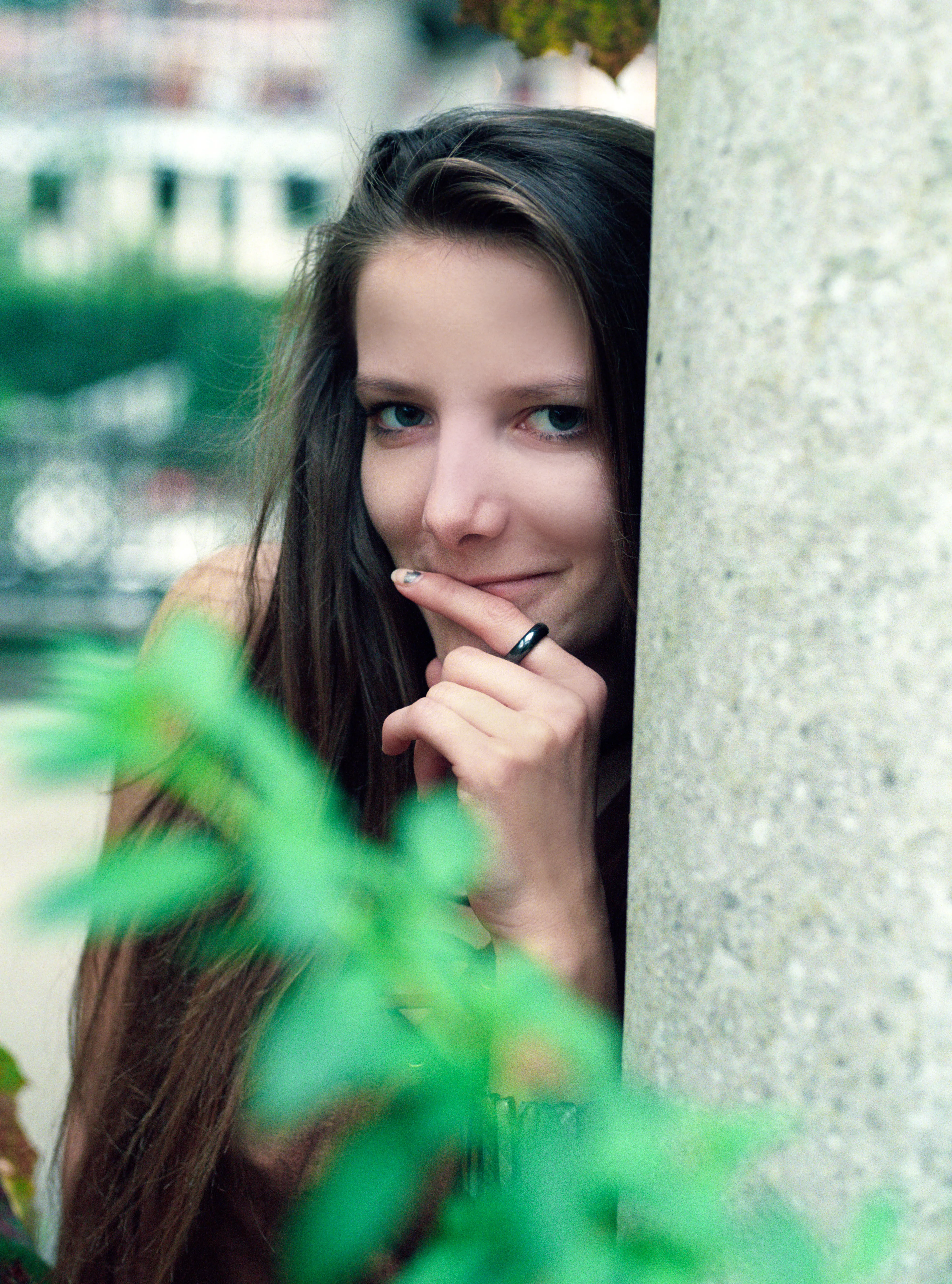 Bella peering around a stone column with a finger to her lips, a green plant blurred in the foreground.
