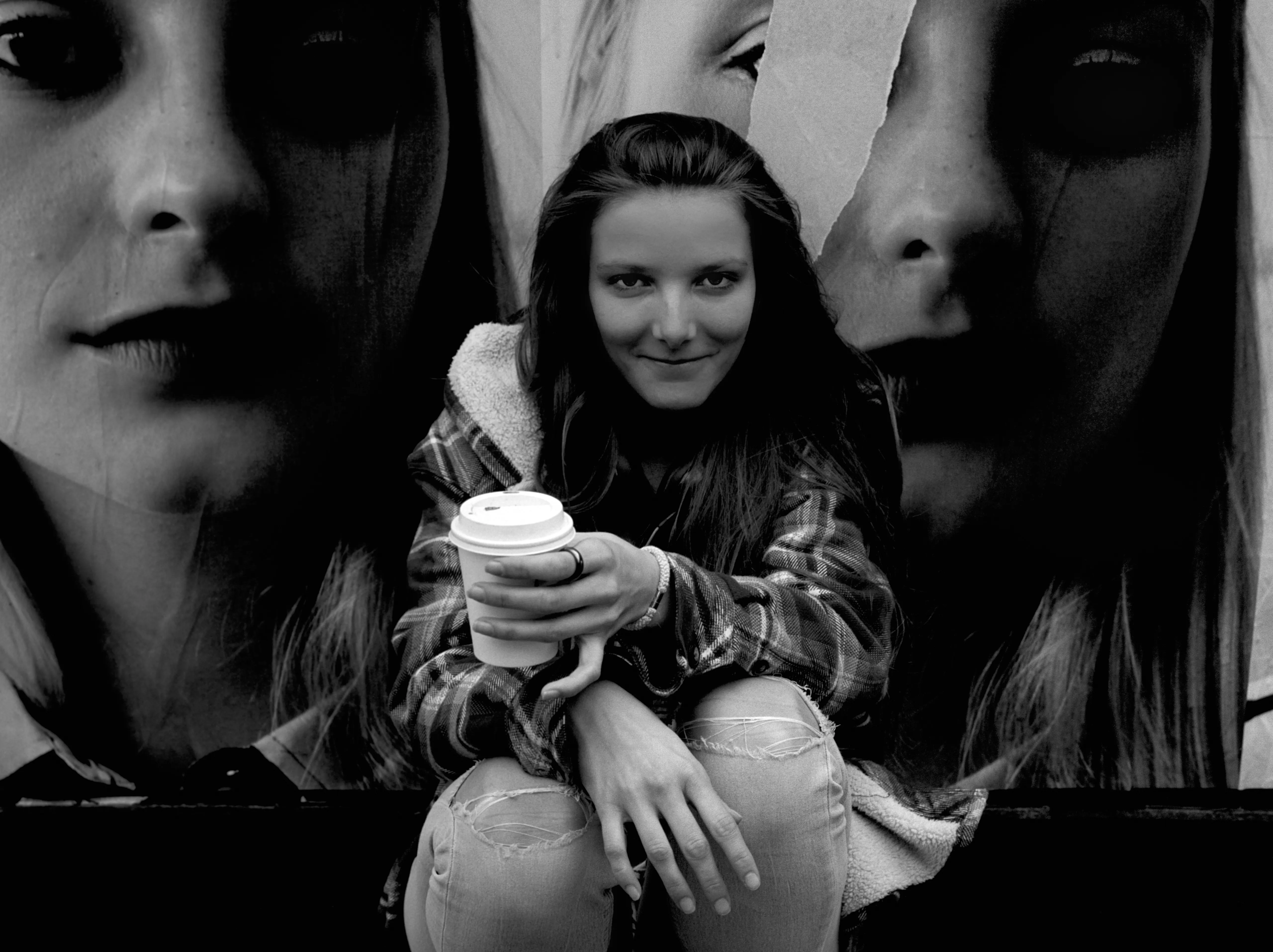 Bella crouched with a coffee cup between two giant pasted faces on a billboard. Black and white.