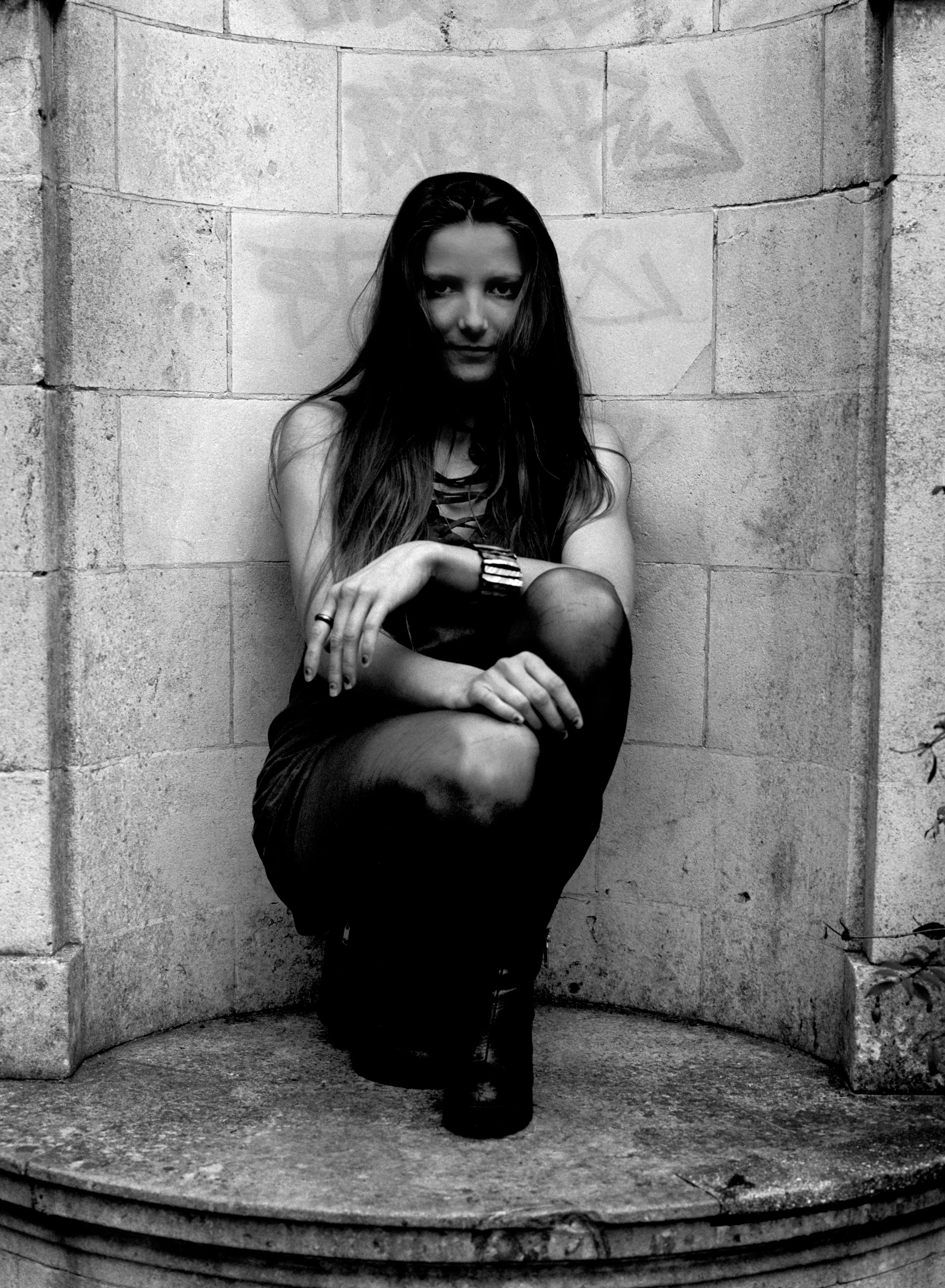 Bella crouched into a curved stone niche, faint graffiti on the wall above. Black and white.