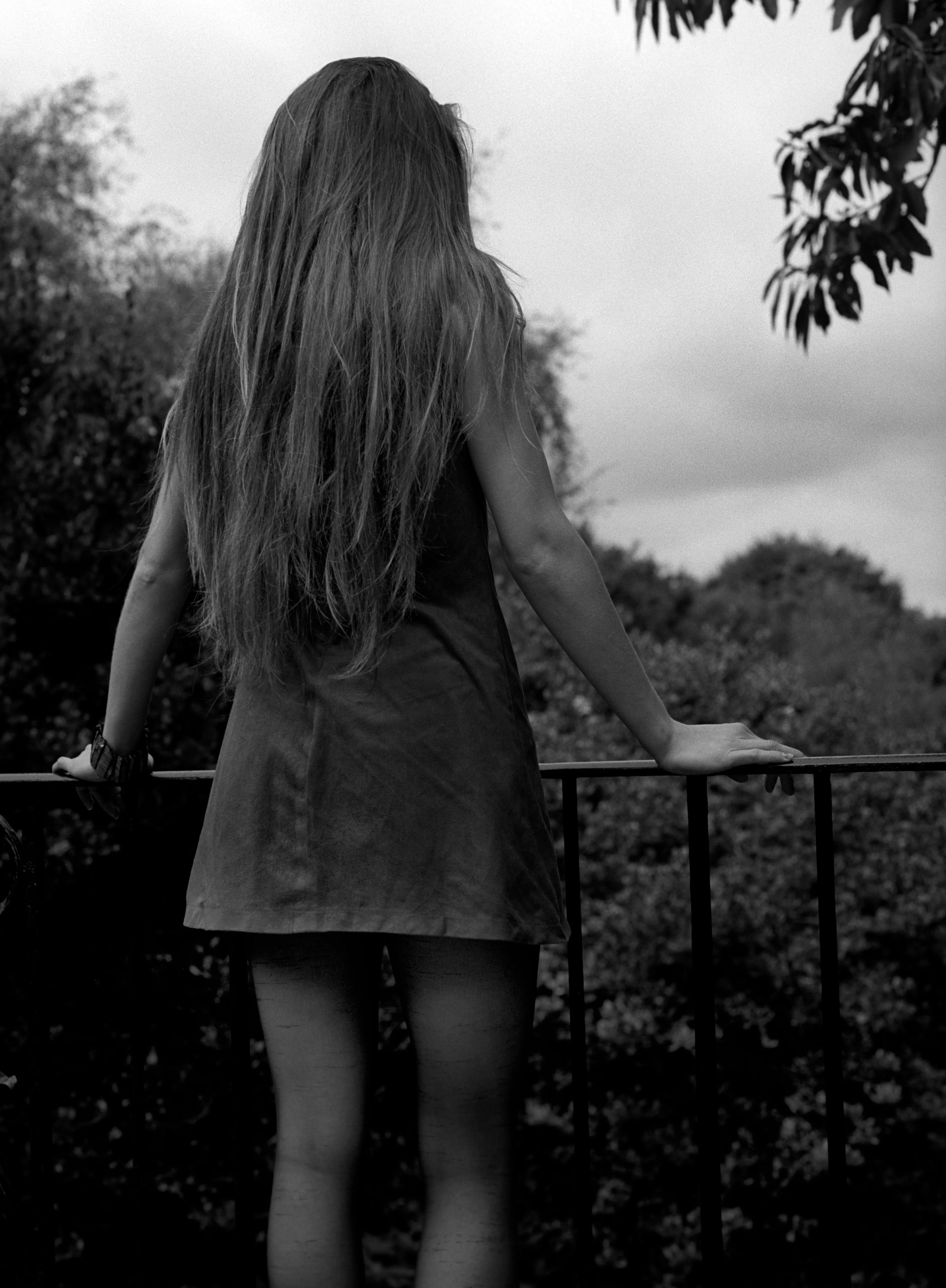 Bella seen from behind at an iron railing, long hair down her back, looking out to a grey sky. Black and white.