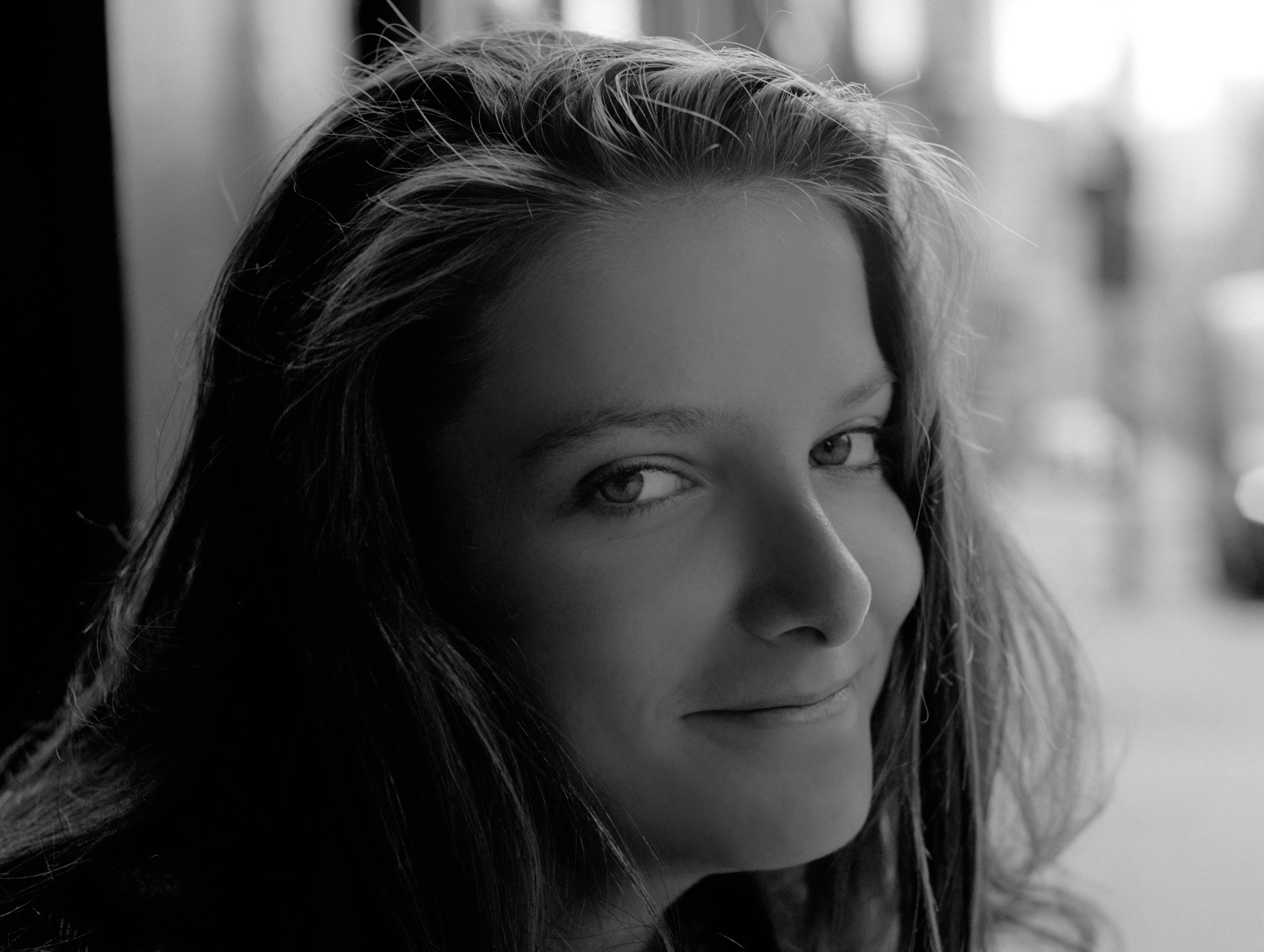 Close portrait of Bella turning to the camera with a faint smile, a blurred street behind. Black and white.
