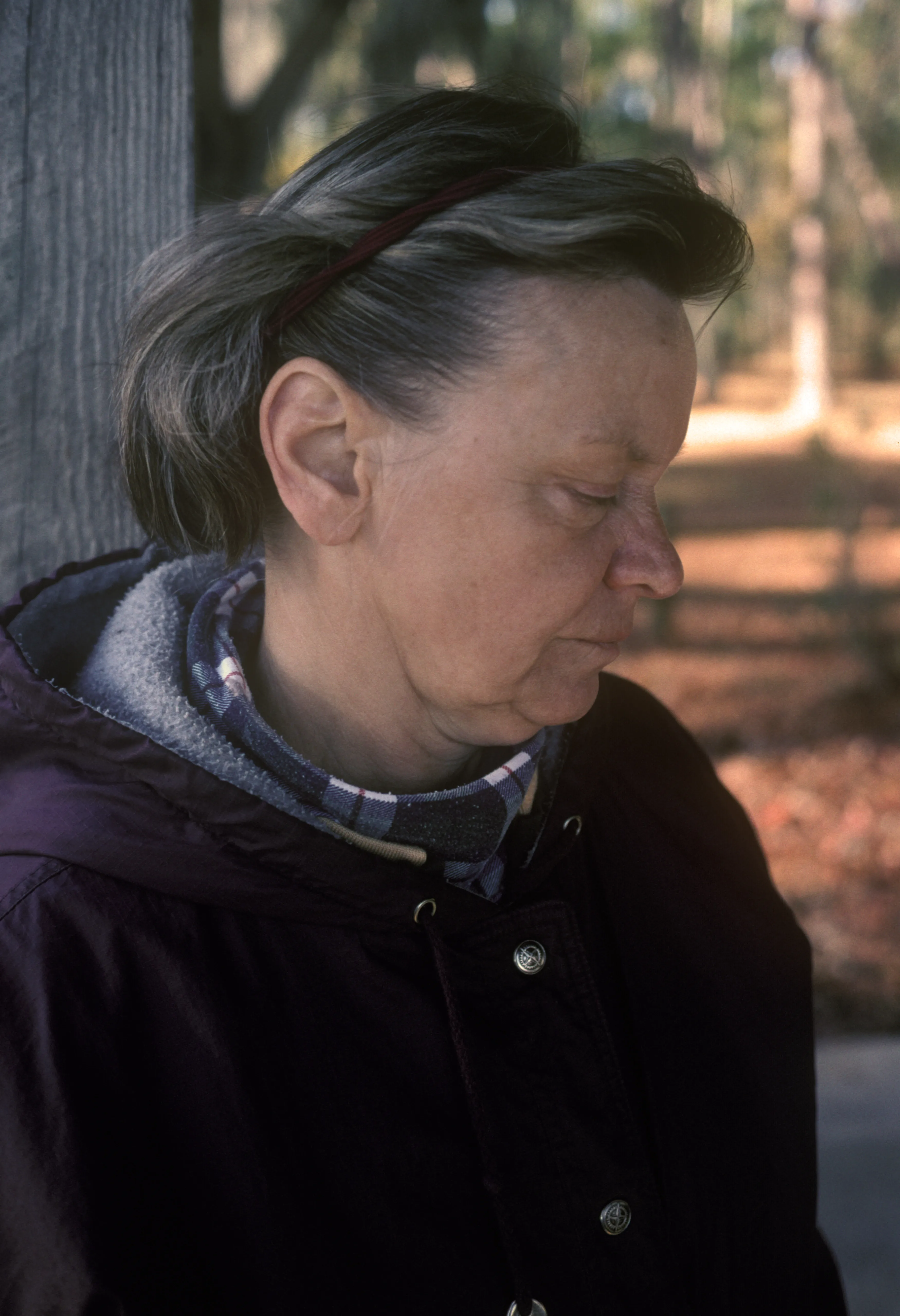 Brenda in profile beside a weathered wooden post, eyes lowered, wearing a dark coat.