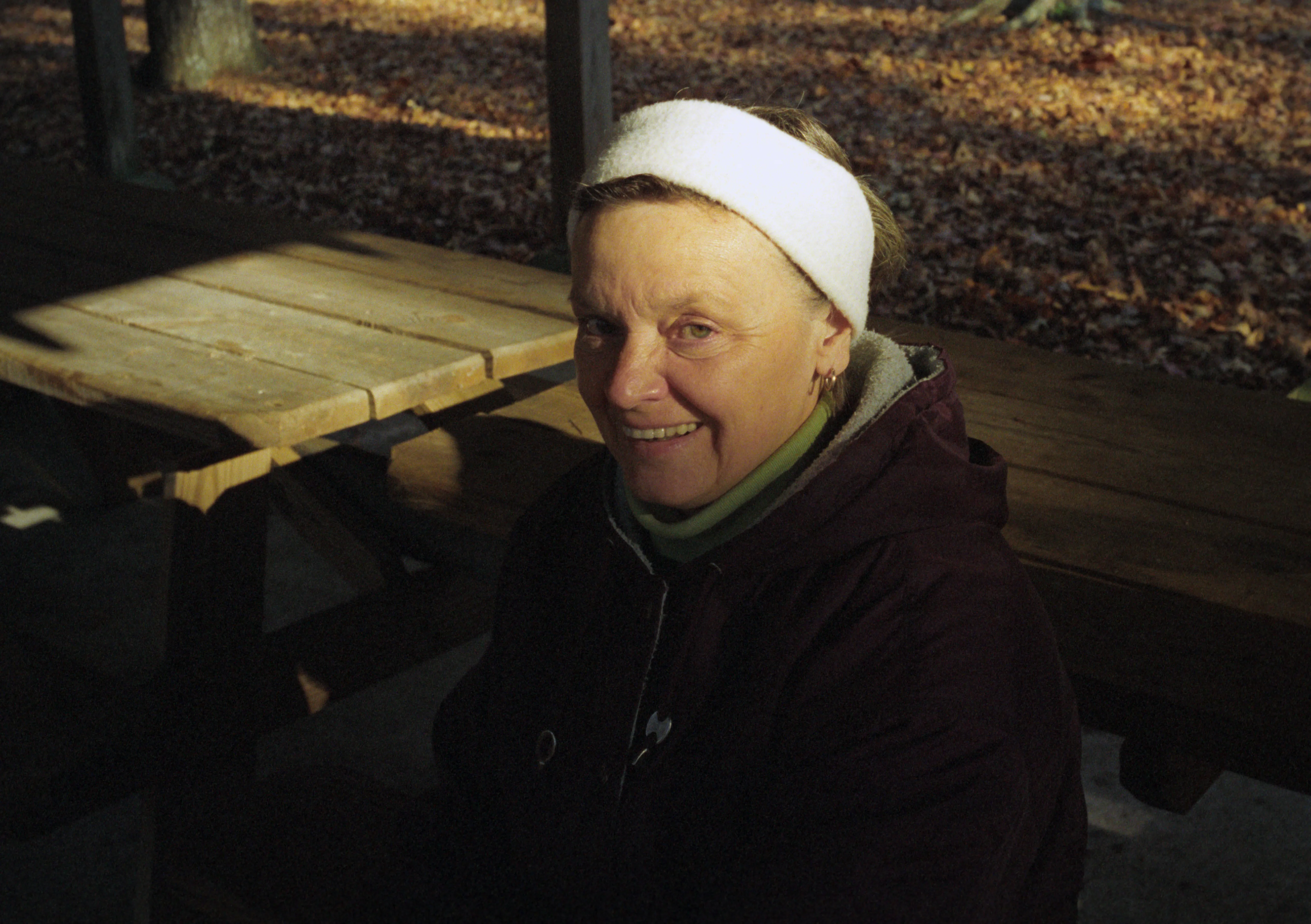 Brenda in a white headscarf and dark coat seated at a wooden picnic table outdoors.