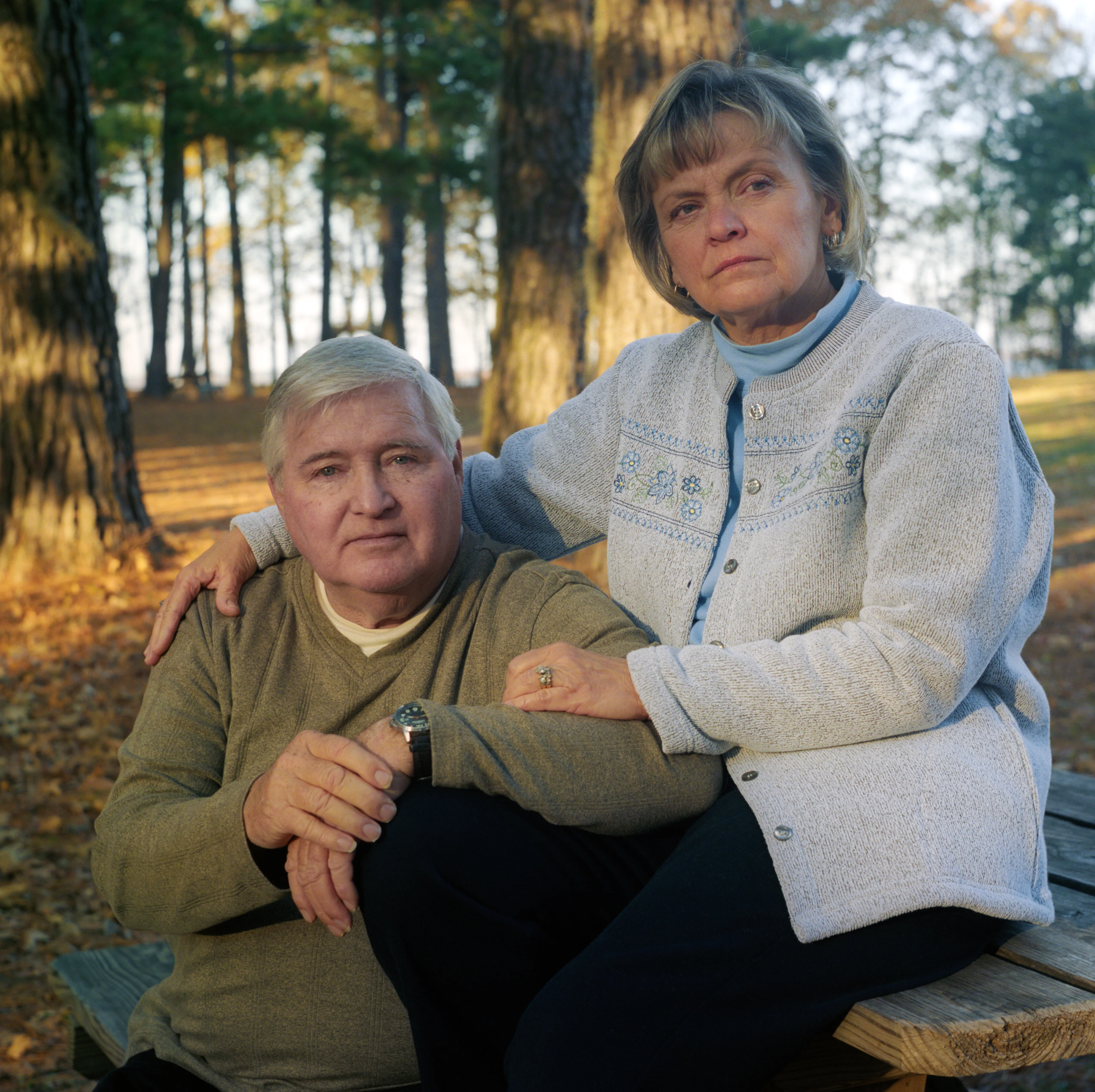 Brenda and her husband Michael seated together on a wooden bench in autumn woodland, her hand resting on his shoulder.