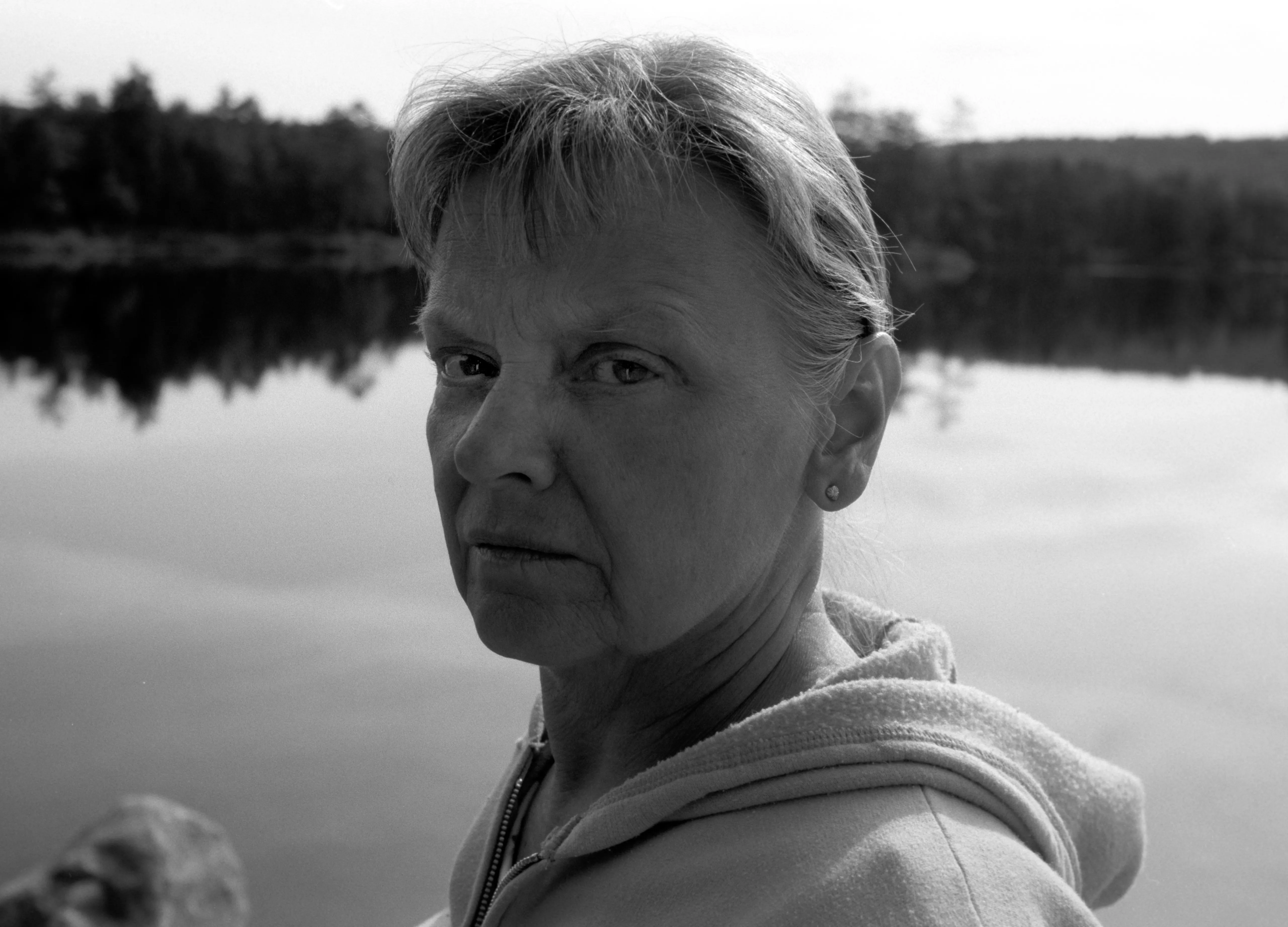 Brenda beside a still lake with a wooded far shore, in a pale hooded top, looking directly at the camera. Black and white.