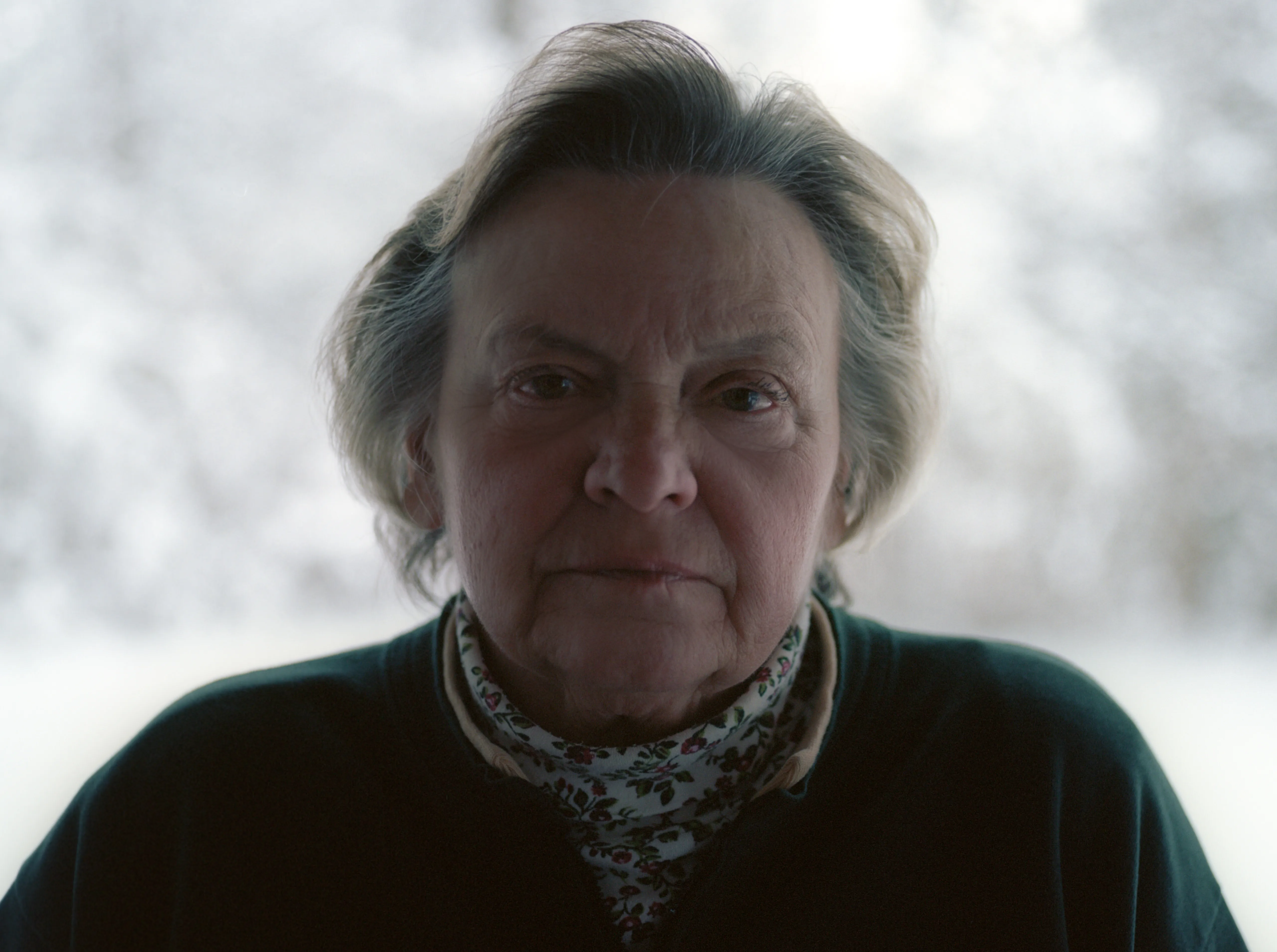 Brenda at the window facing the camera, snow-covered trees behind her, in a dark top and floral scarf.