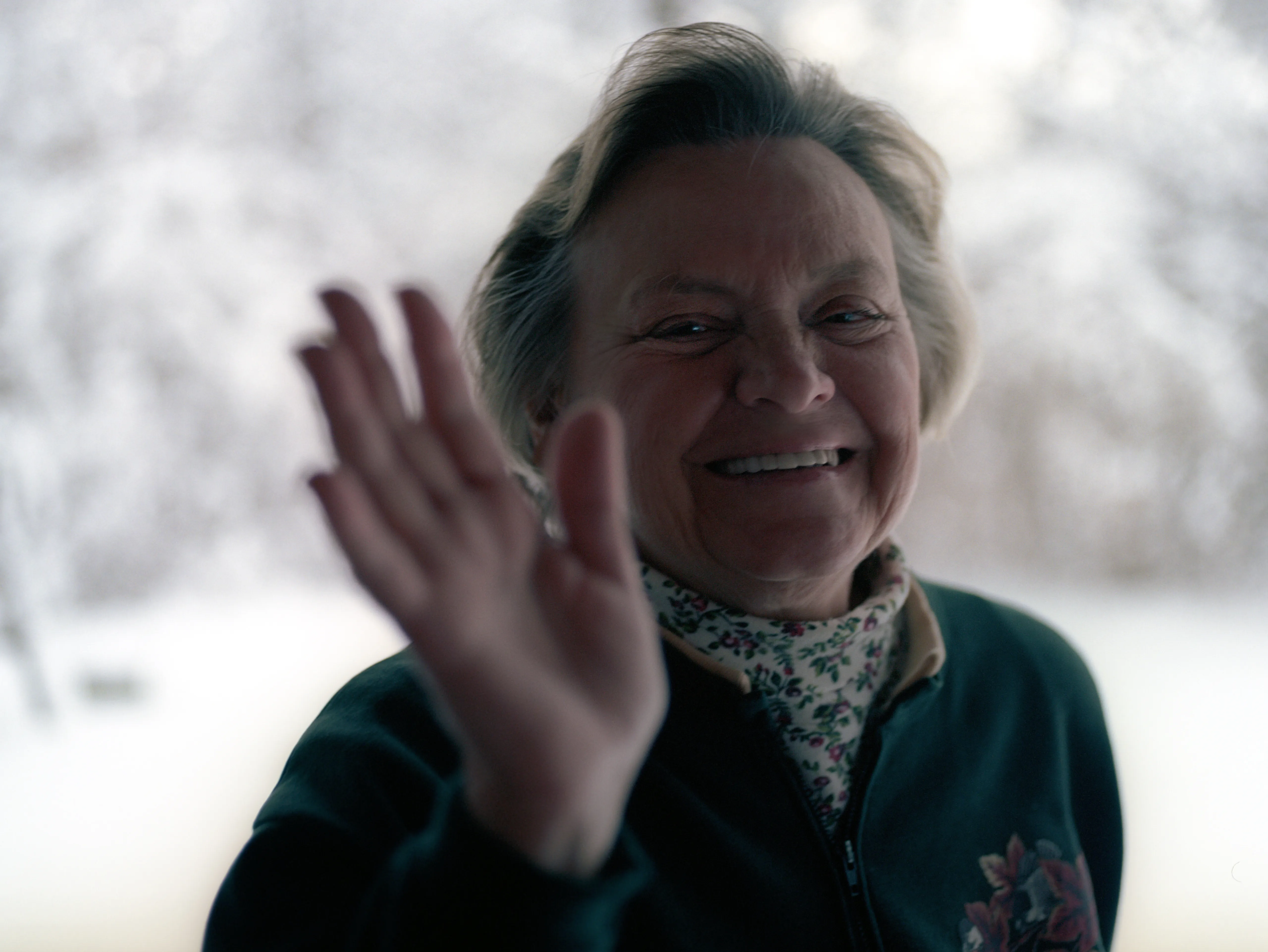 Brenda at the window, one hand raised in a wave, smiling, snow outside.