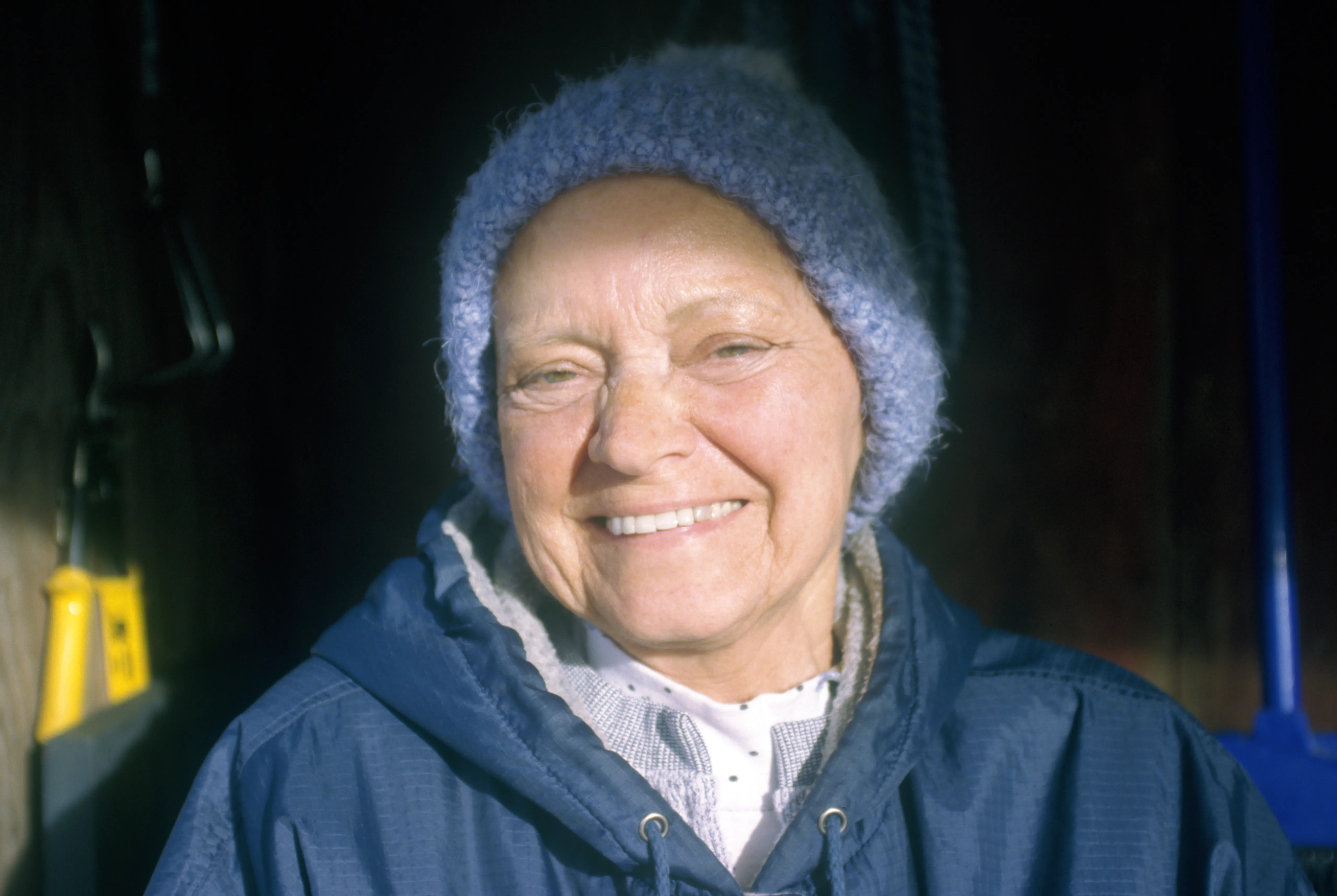Brenda in a pale knitted hat, lit from the side against a dark interior, laughing.