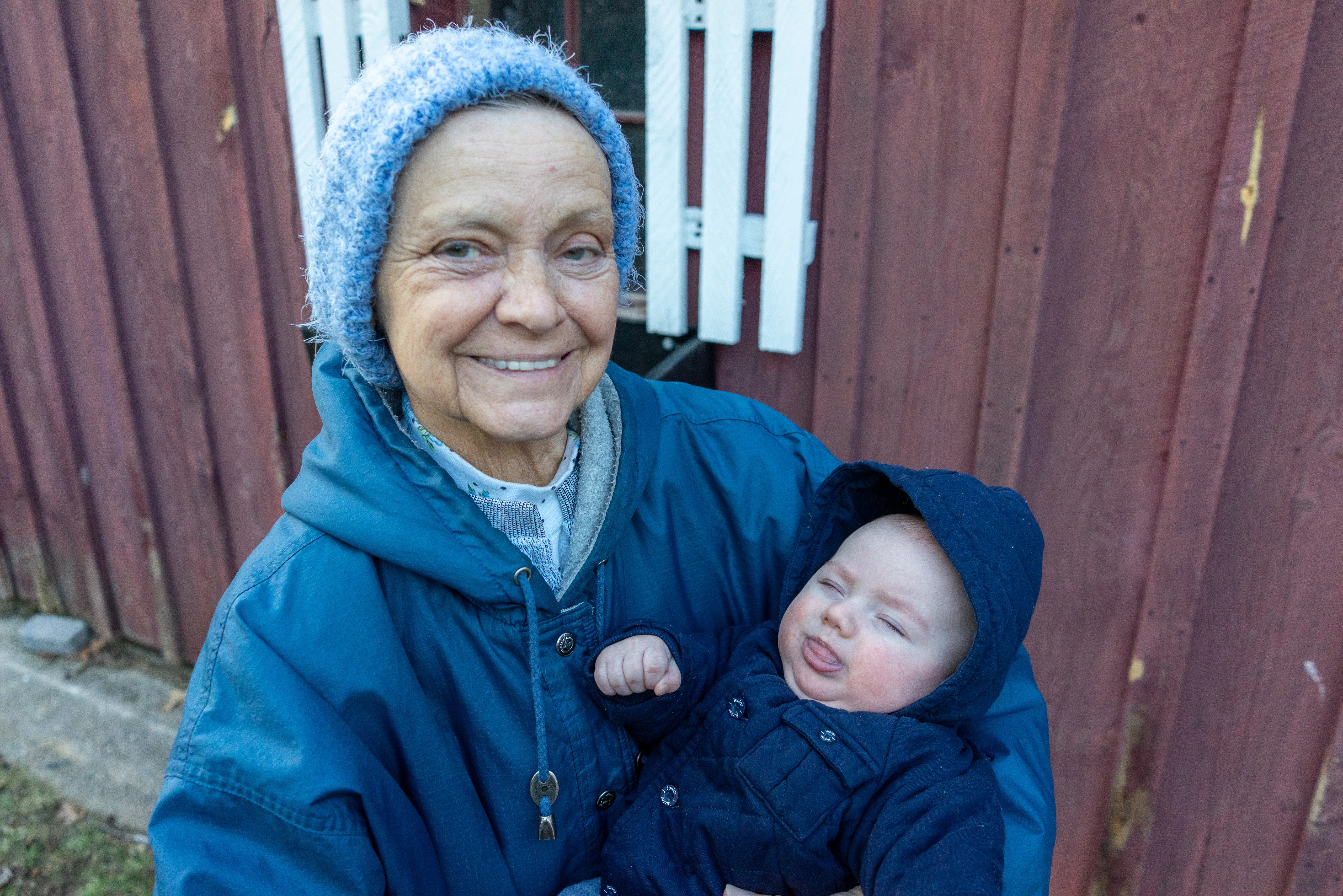 Brenda in a knitted hat and blue coat holding her infant grandson, standing before red-painted barn boards; a smile.