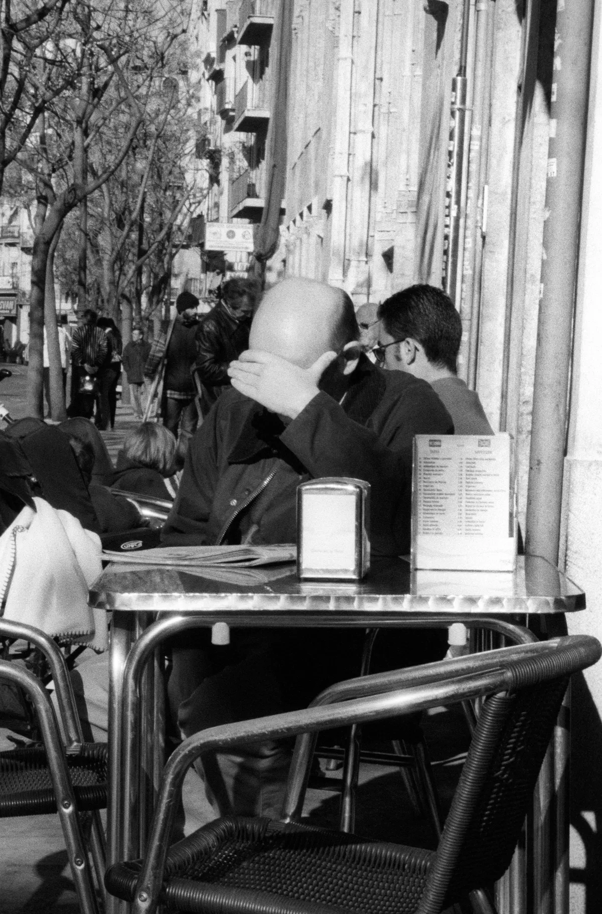 A bald man at a café terrace table, hand screening his face from the light.