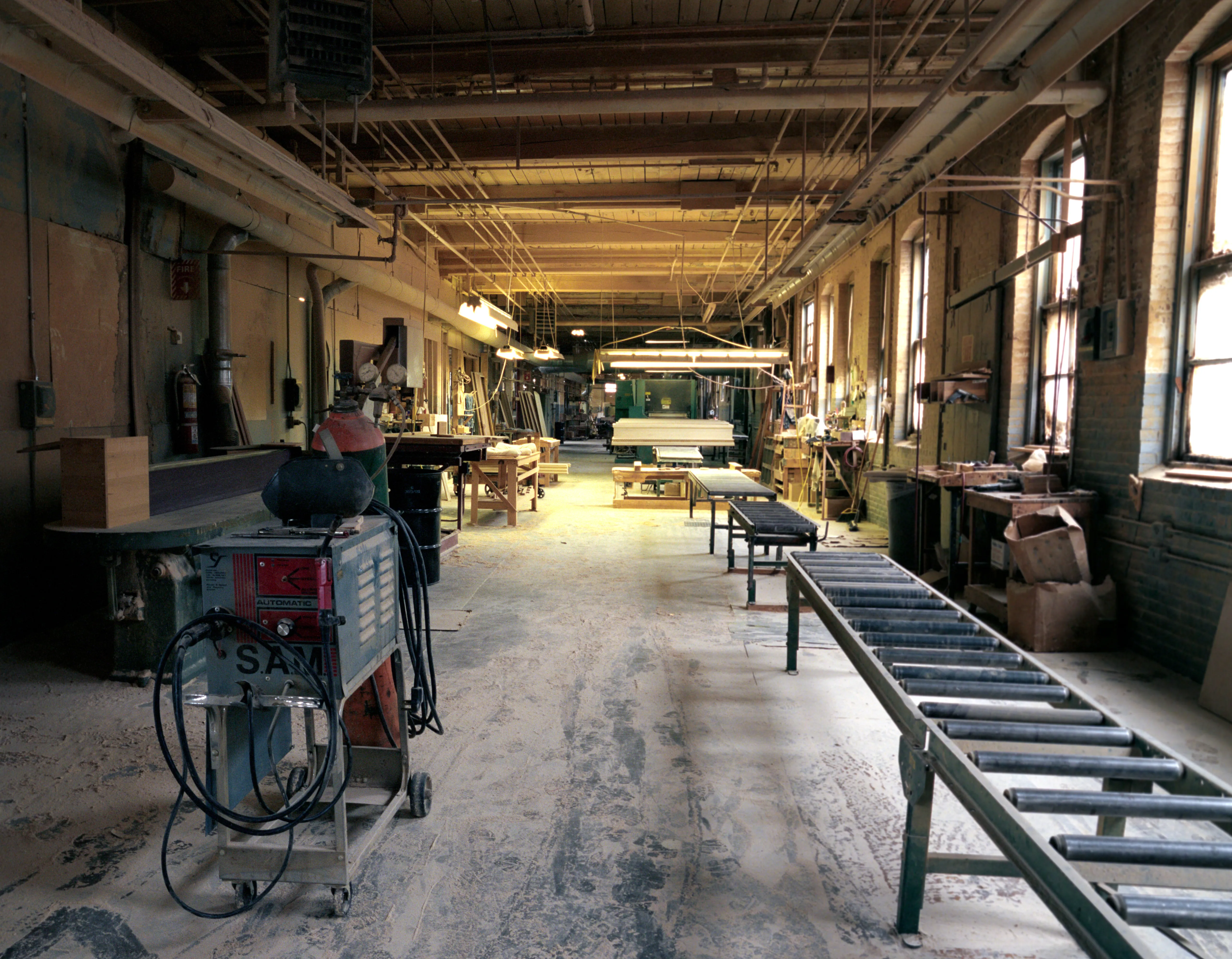 Down an aisle deep in wood dust, a pale casket rests on trestles midway; at left a cart of coiled hoses and a gas bottle, at right steel rollers under arched windows.