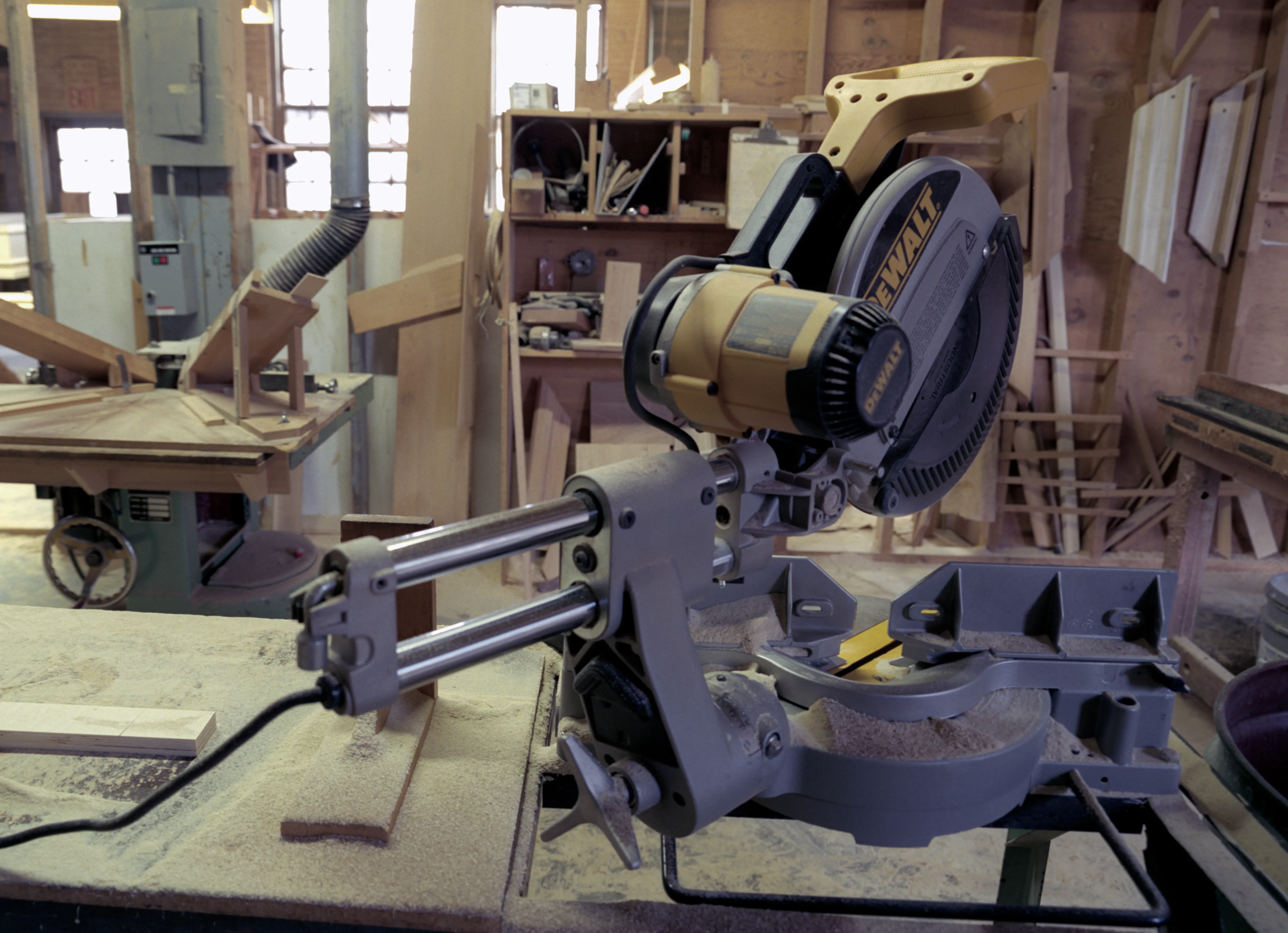 A yellow-and-black sliding mitre saw fills the foreground on a bench under fine dust, offcuts beside it; behind, a cast-iron machine with a handwheel, tool shelves and windows in a timber-framed shop.