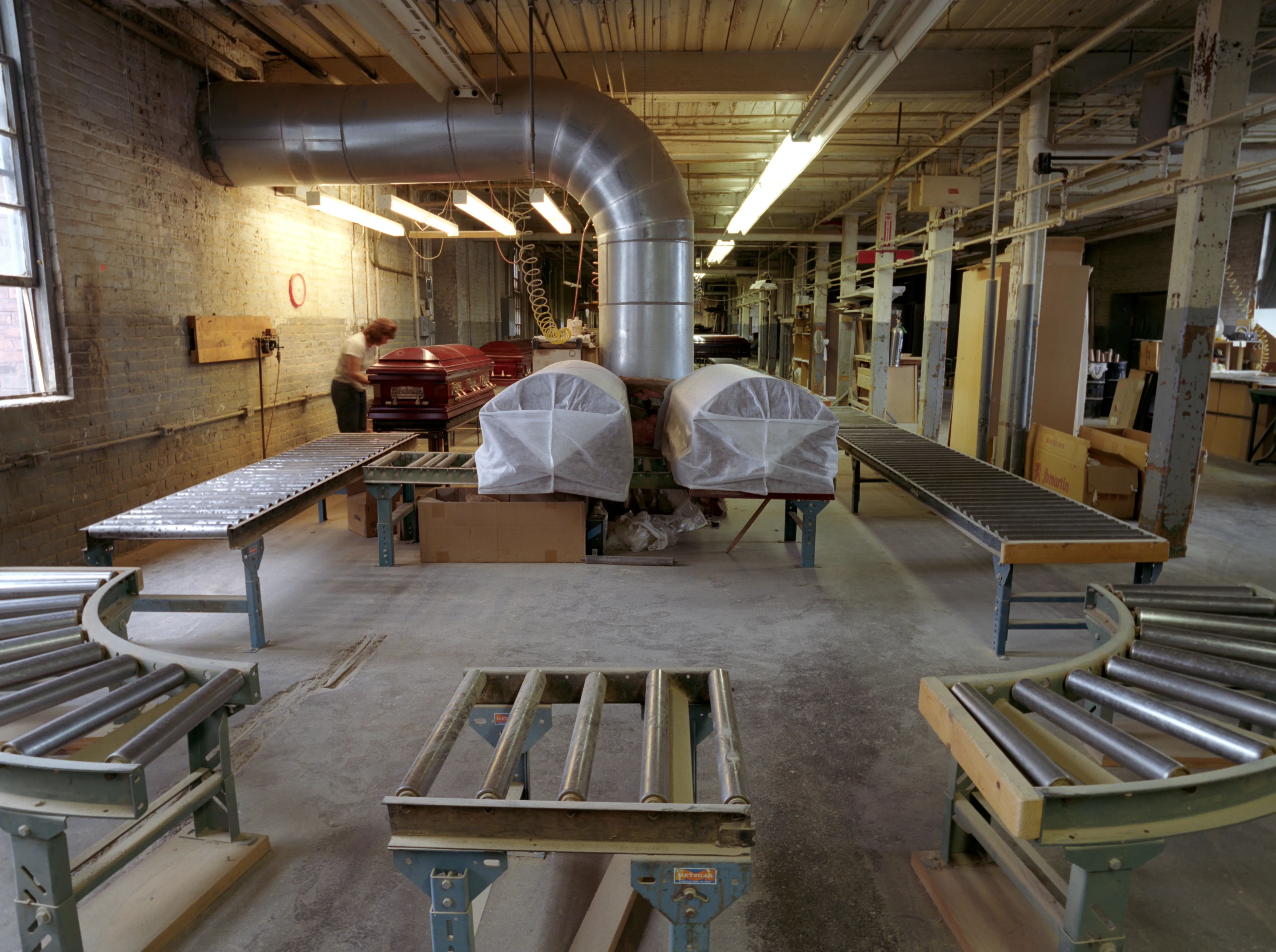 Shrouded in white covers, two machines stand beneath a silver extraction duct; roller conveyors curve across the concrete, and by the wall someone in a pale top bends over a red casket.