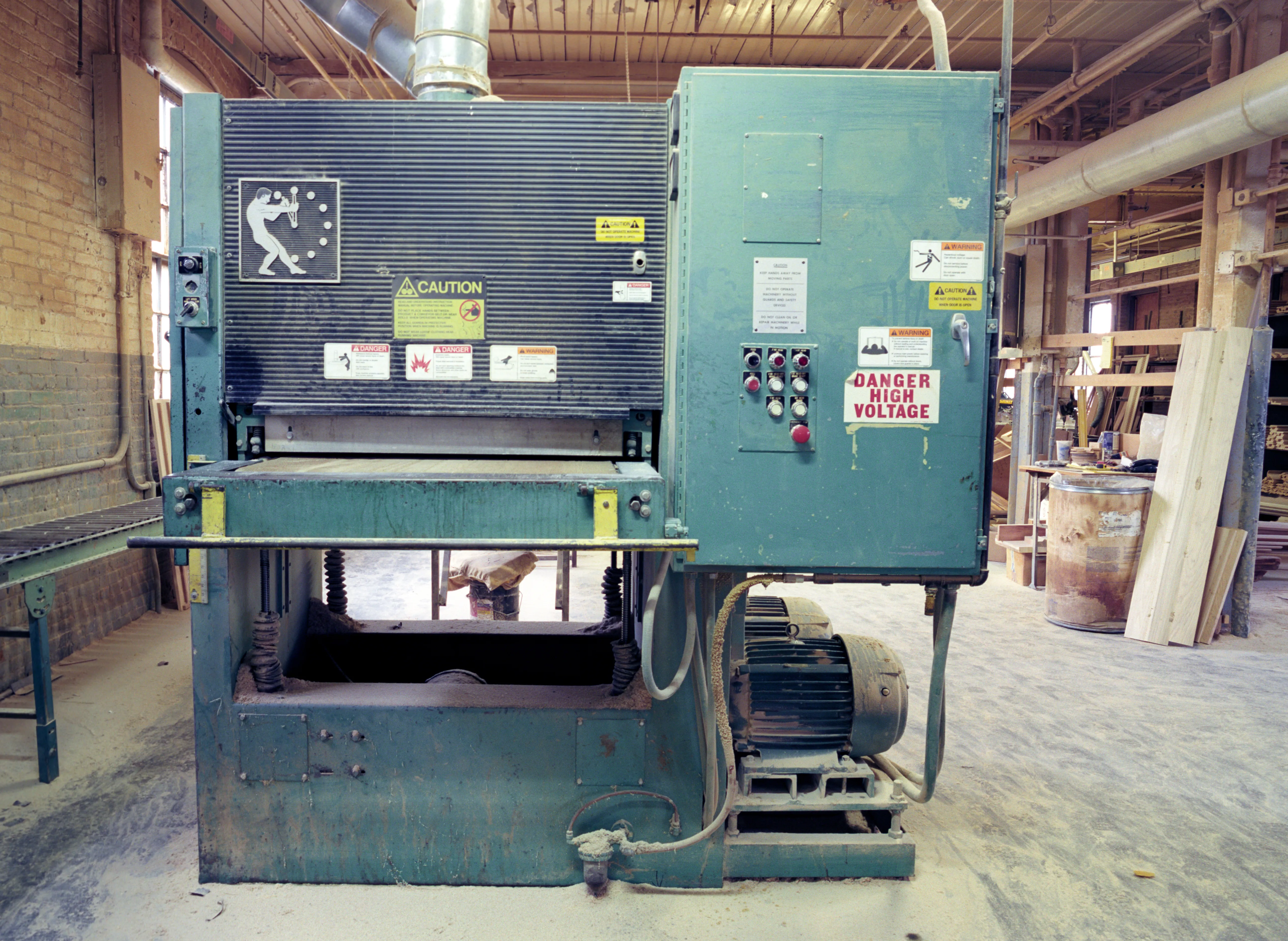 Head-on, a green woodworking machine carries a warning pictogram and caution panel, beside it a switch cabinet of push buttons and a high-voltage placard; dust over the floor, boards leaning at right.
