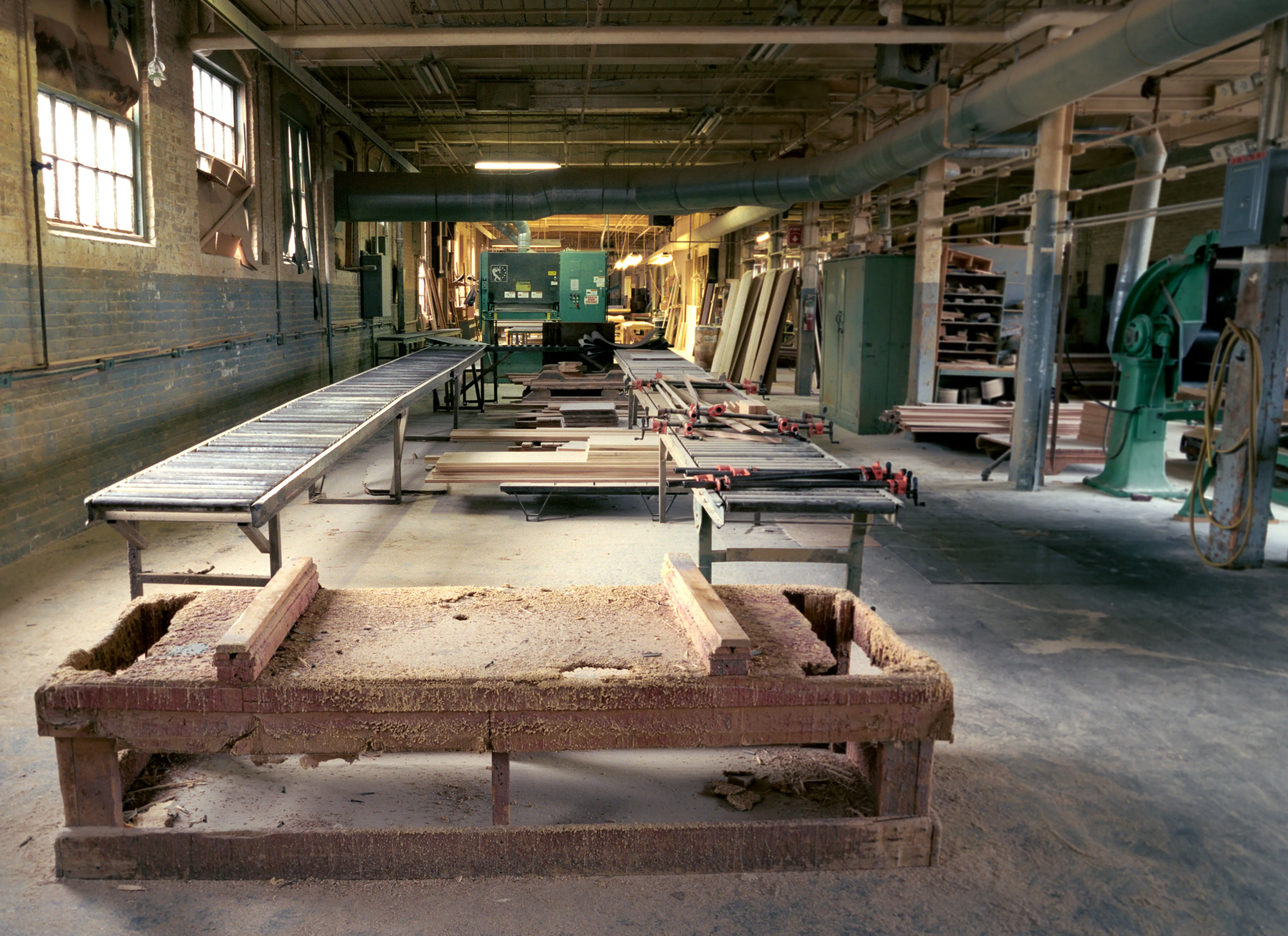 The nearest bench lies buried under sawdust; behind it a roller table and boards gripped in long red clamps run back towards cabinets and racking, daylight falling from two tall windows.