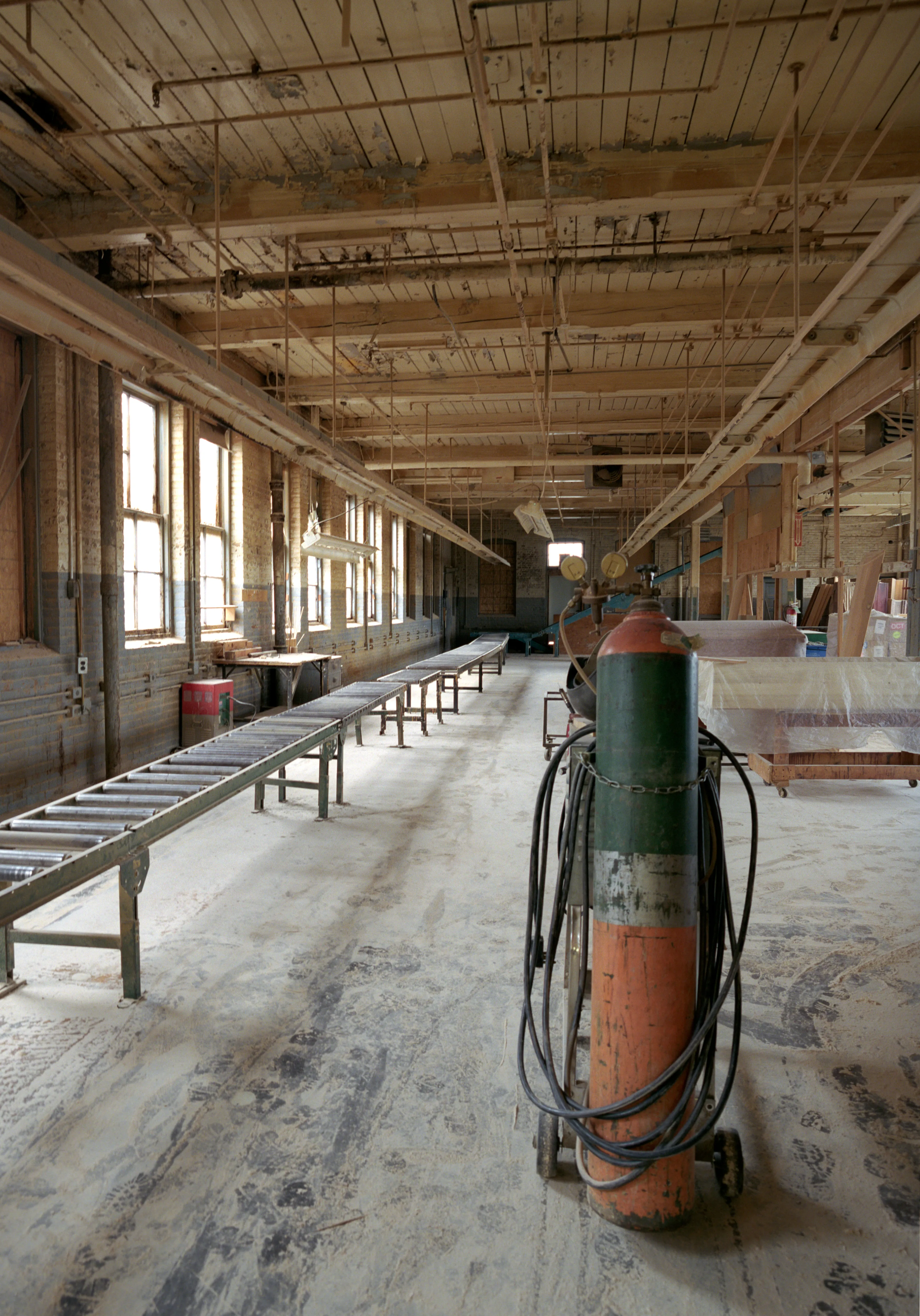 Black hoses loop round a gas cylinder chained to a wheeled trolley on scuffed concrete; a conveyor on trestle legs runs down the sunlit window wall, plastic-wrapped caskets on carts at right.