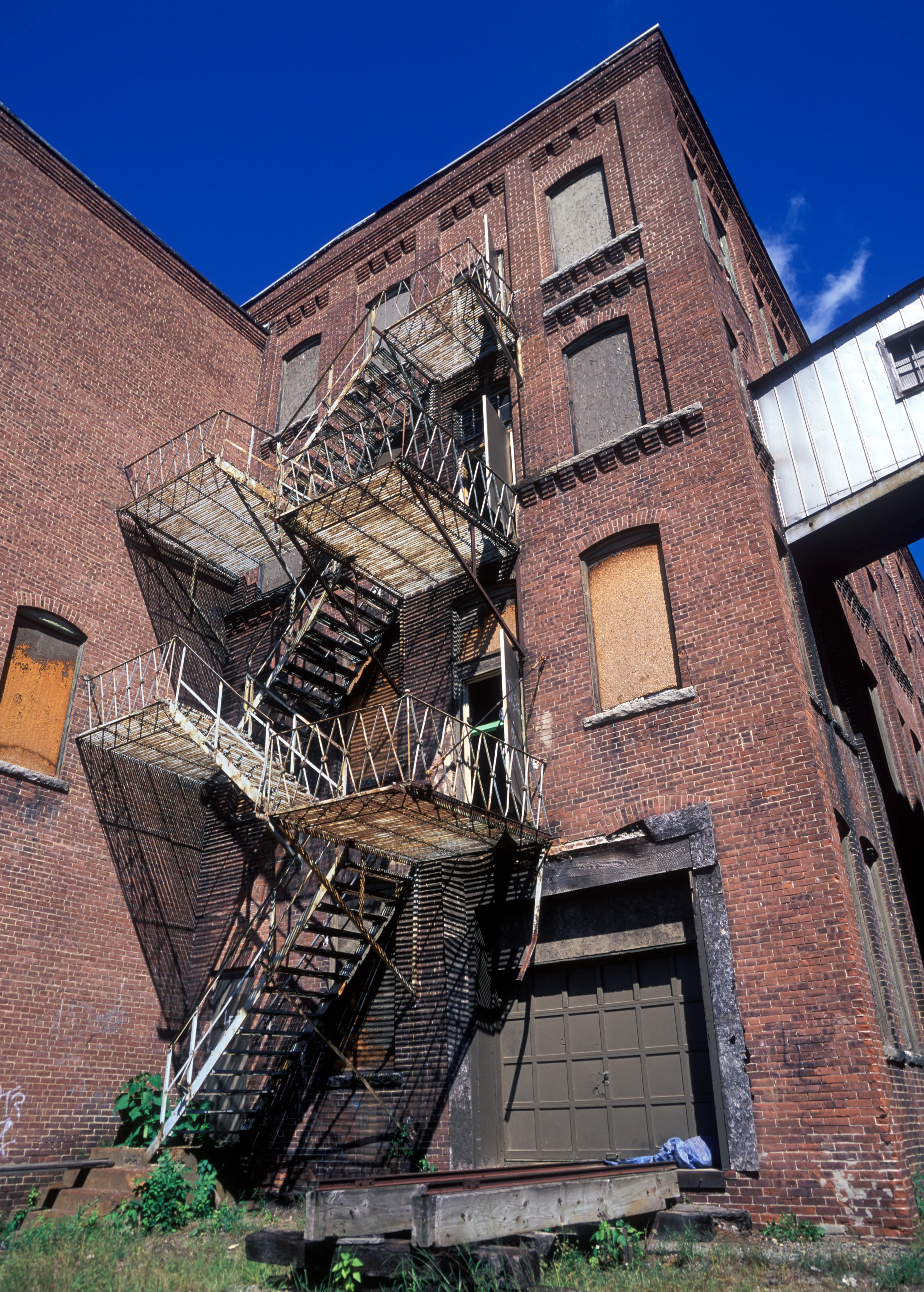 Zigzagging down the corner of a four-storey brick mill against saturated blue sky, a rusting fire escape drops its timber landings almost to the ground; boarded windows, an olive loading door below.