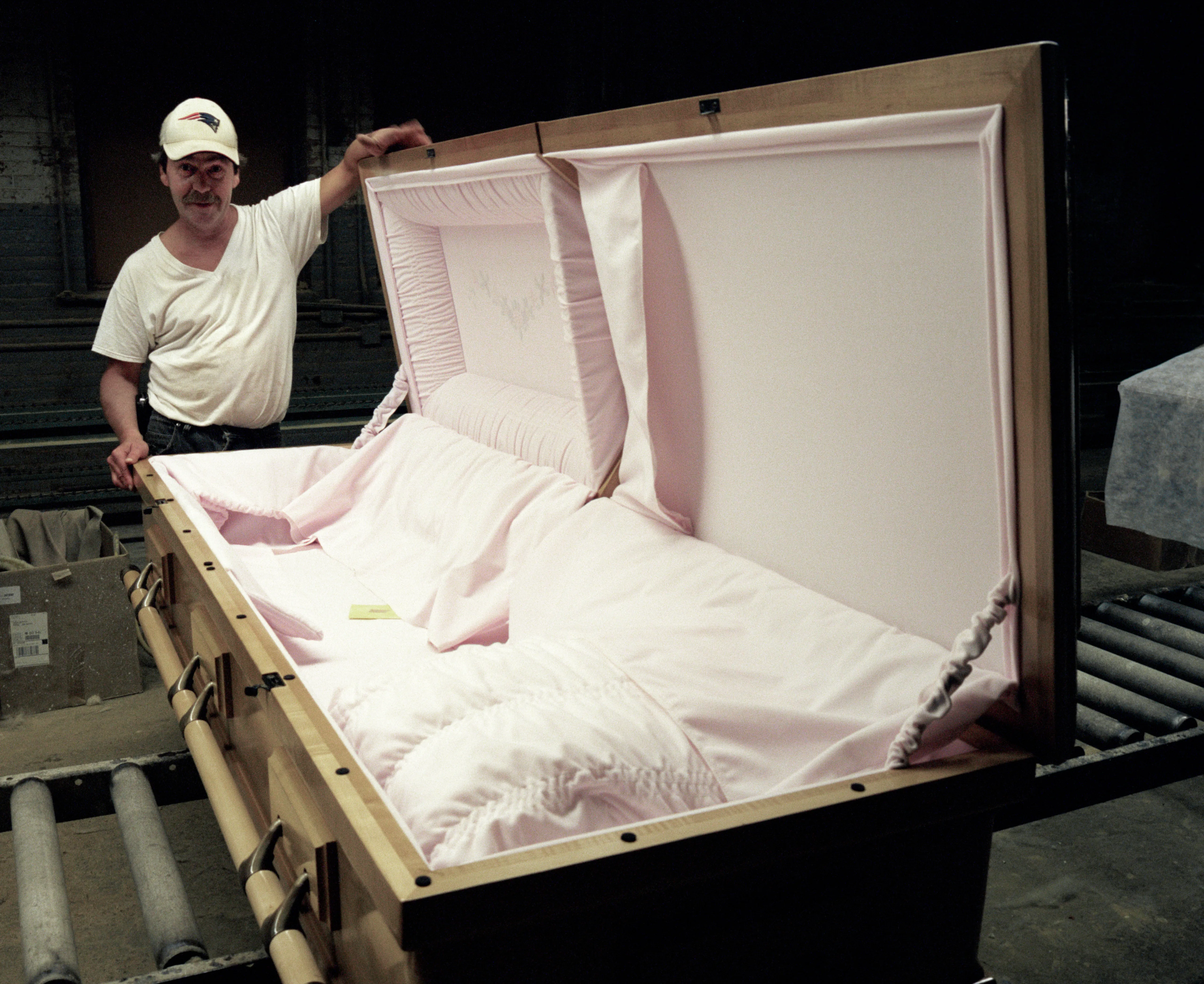 At the head end stands a person in white cap and T-shirt, gripping the raised lid of a casket lined in pale pink satin; it rests on rollers, the room behind black.