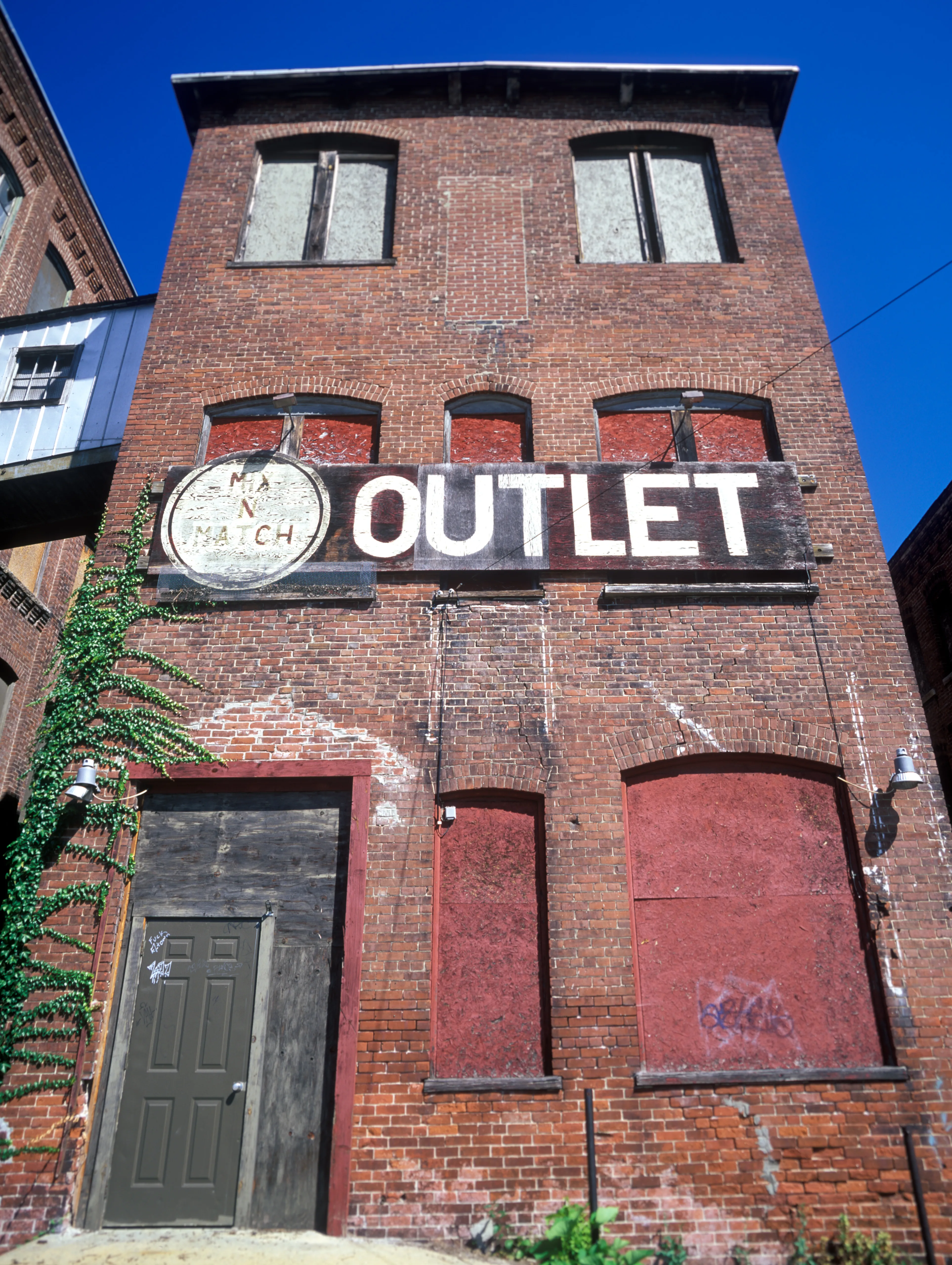 OUTLET in white capitals spans a board bolted across the middle storey of a weathered red-brick front; boarded openings above, red-painted arches below, a panelled door and climbing ivy at left.