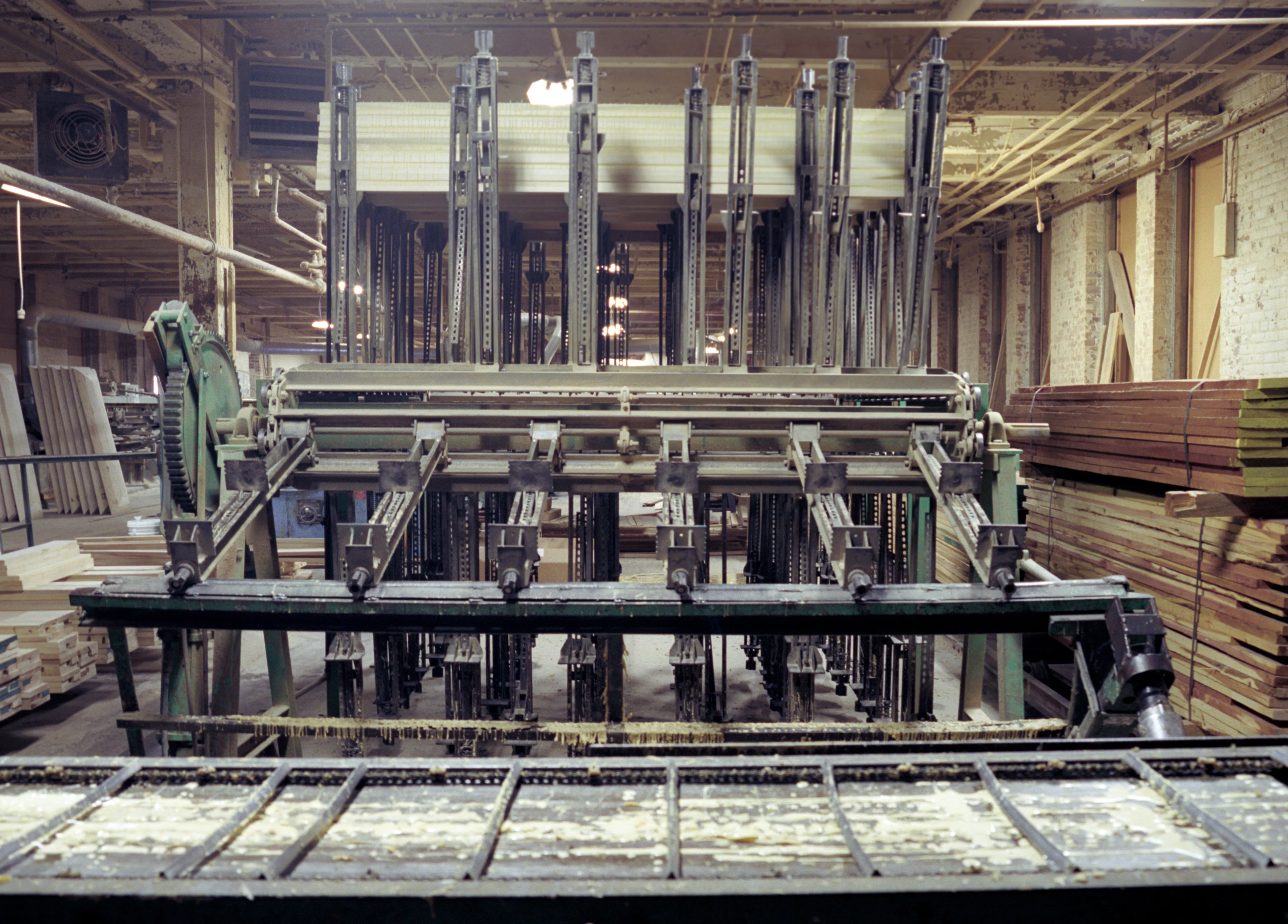 Ranks of vertical steel clamps rise from a green-framed machine towards the beamed ceiling; its wide slatted bed, caked with dried glue, crosses the bottom of the frame, sawn boards stacked right.