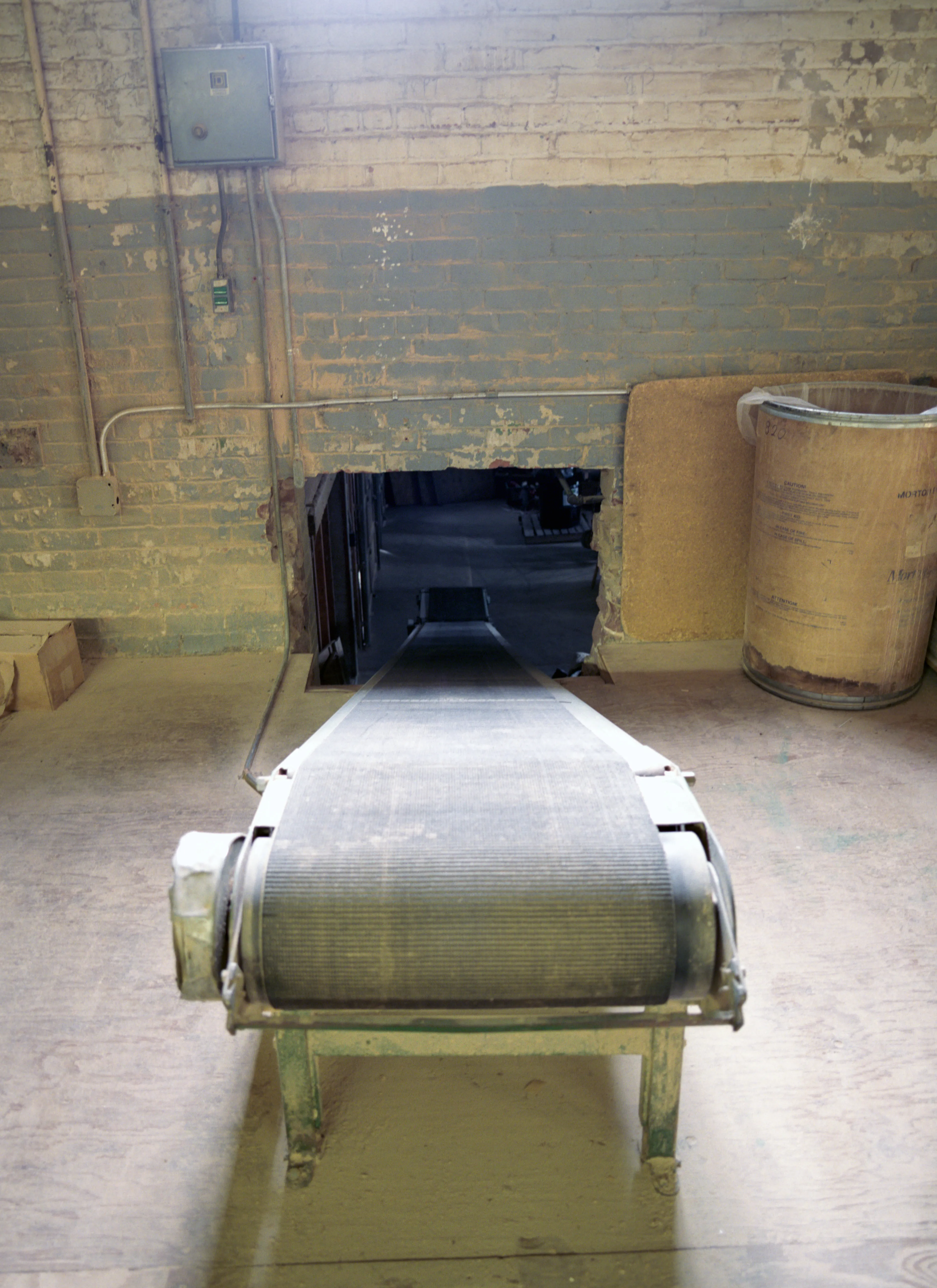 Where a rough hole breaks the peeling brick wall, a fabric conveyor belt on a green-painted frame runs away from the camera into unlit space; a fibre drum at right.