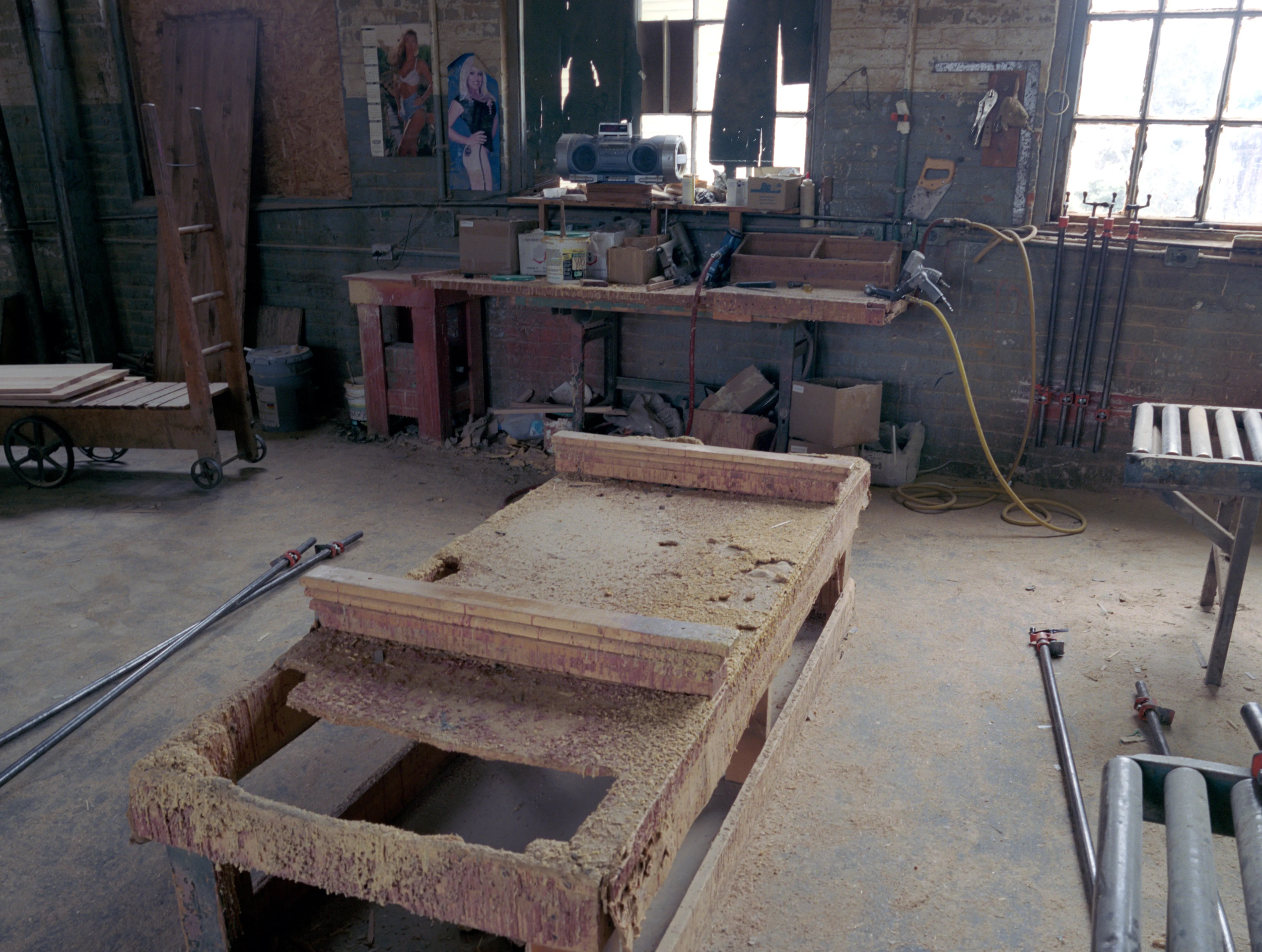 Sawdust crusts the top and overhanging edges of a long workbench that angles in from the corner; past it, steel clamps on the concrete, a cluttered second bench, pin-up posters, bright windows.