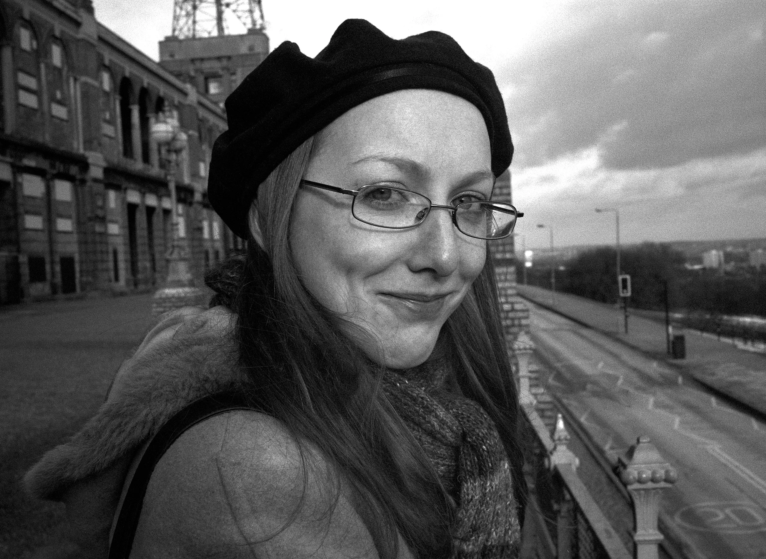 Catherine in a beret and glasses on a terrace, a mast and empty road behind.
