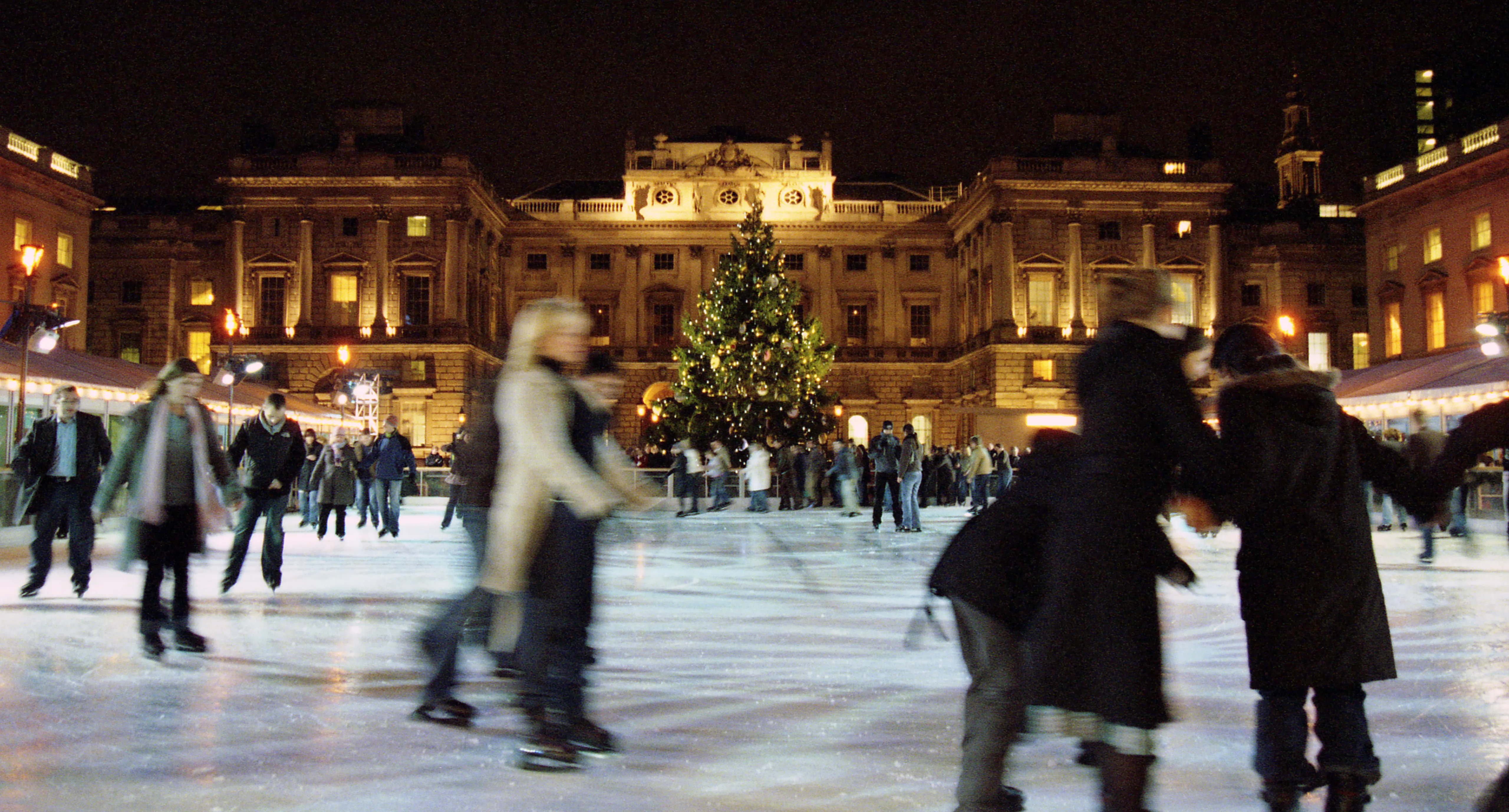 Skaters blurred in motion on the open-air rink at Somerset House at night, a lit Christmas tree and the floodlit façade behind.