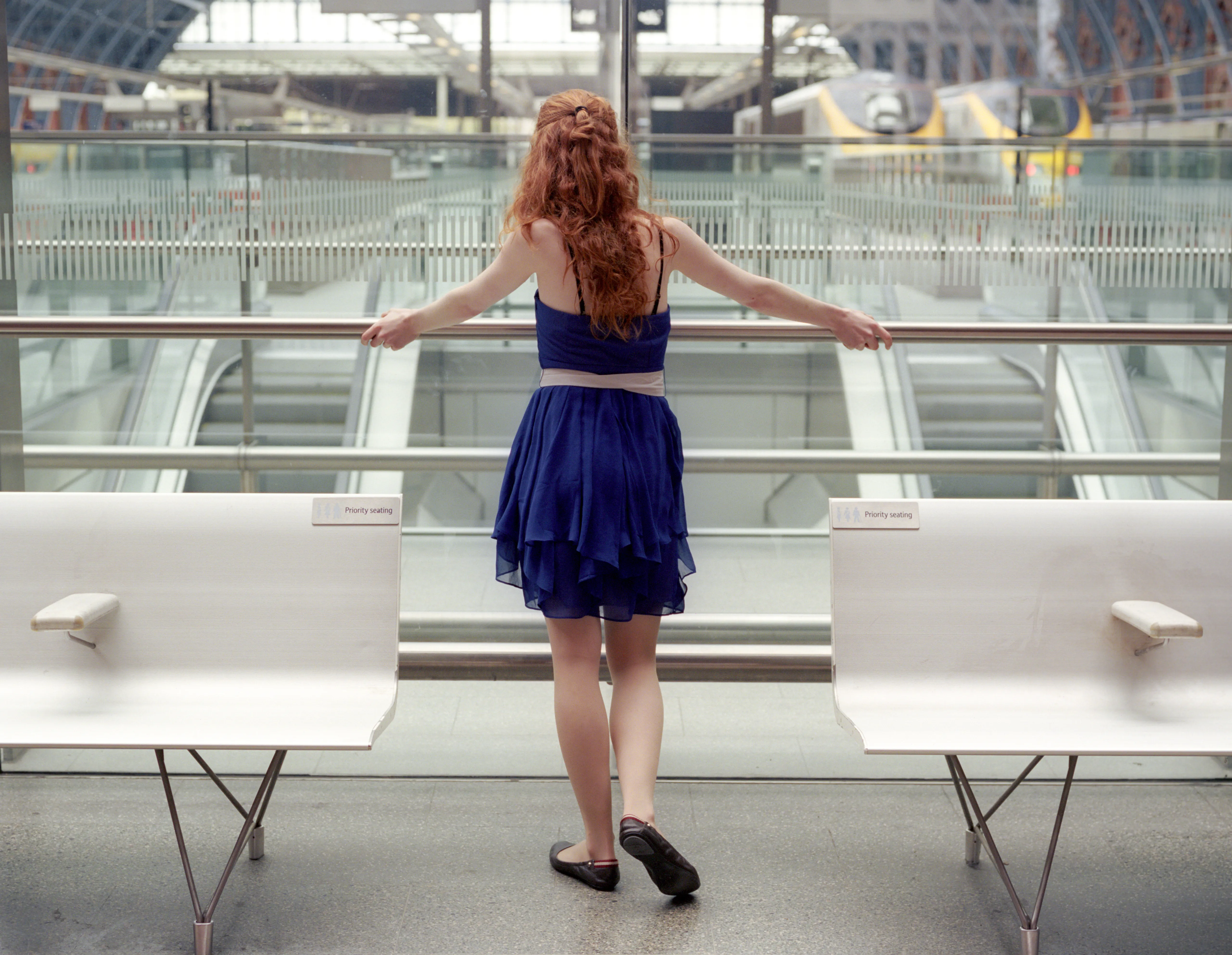 Clélia stands at a glass balustrade inside St Pancras, her back to the camera in a blue tiered dress, red hair down her back, facing the platforms below.
