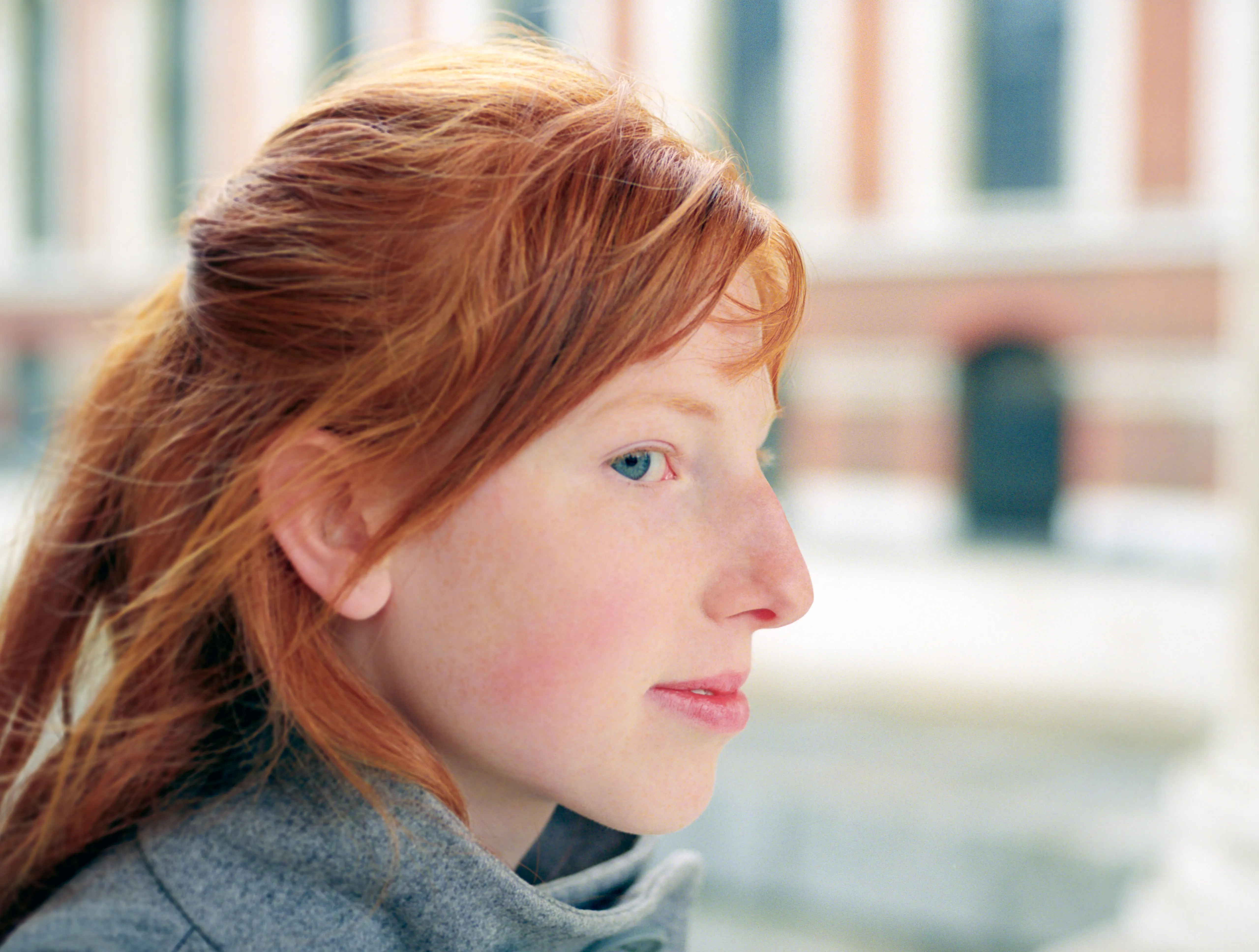 A side profile of Clélia, red hair falling across her face, a blurred building behind her.
