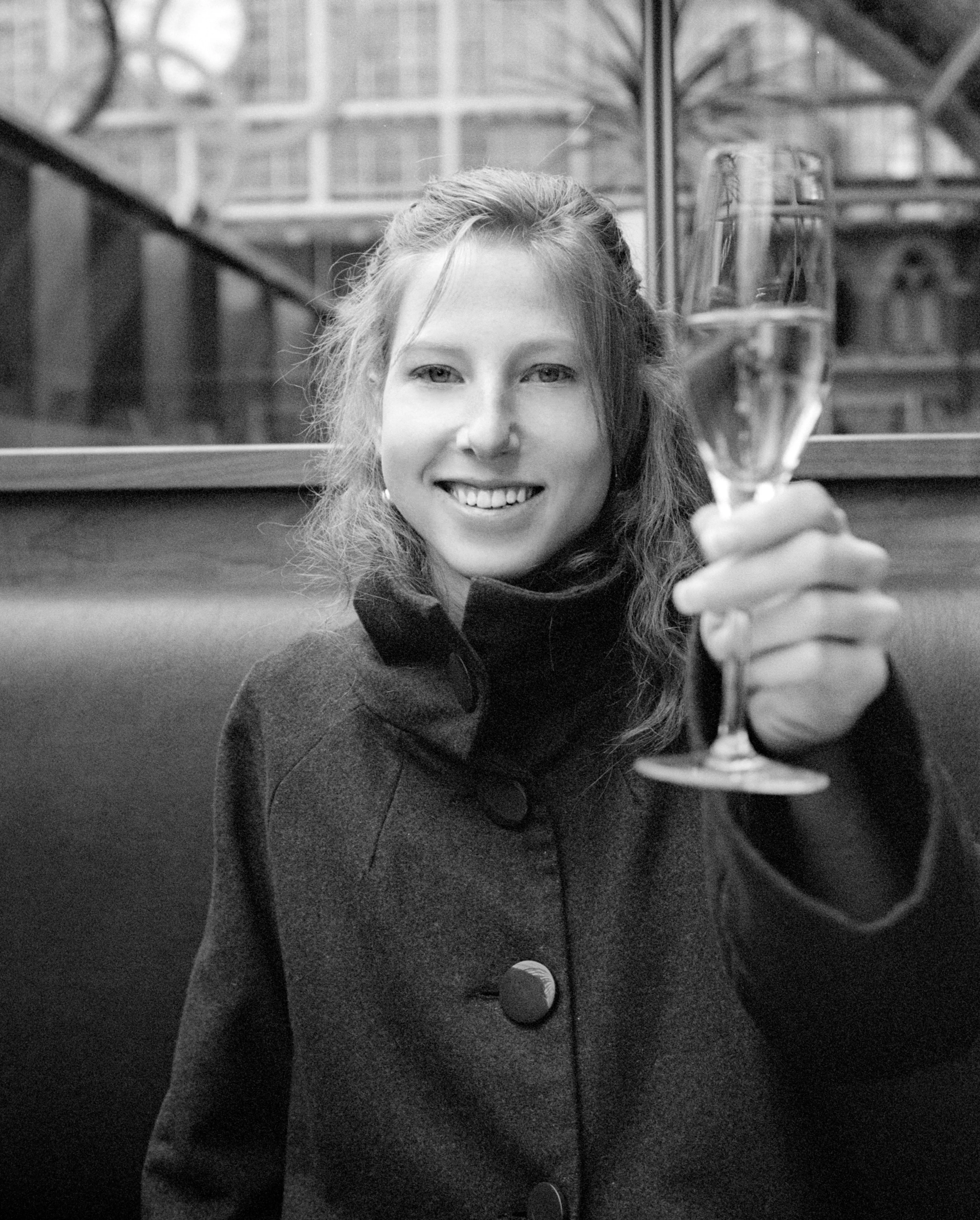 In black and white, Clélia smiles and raises a champagne flute, in a dark high-collared coat.