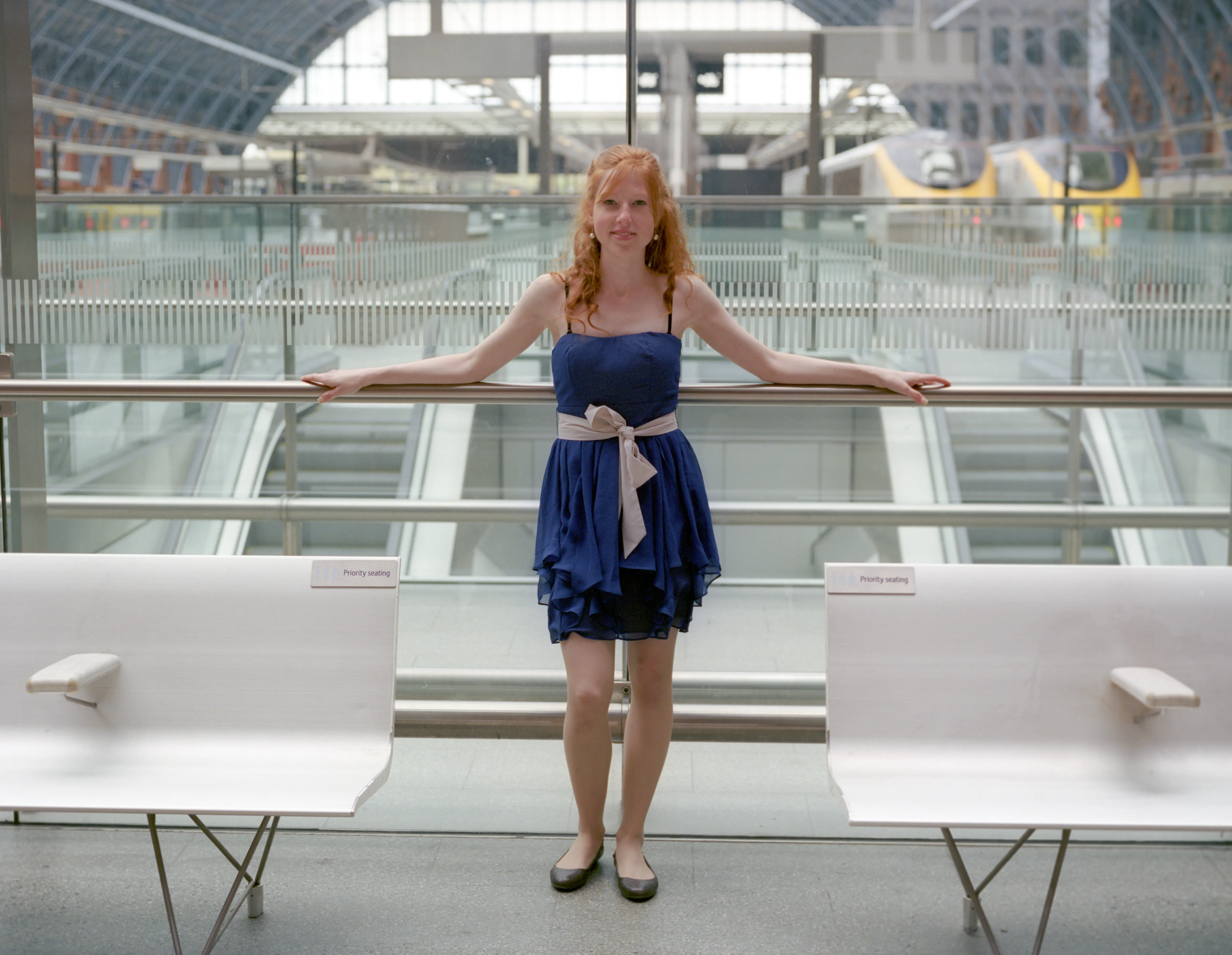 Clélia leans back against the glass balustrade at St Pancras, facing the camera with both arms along the rail, in a blue dress tied at the waist.