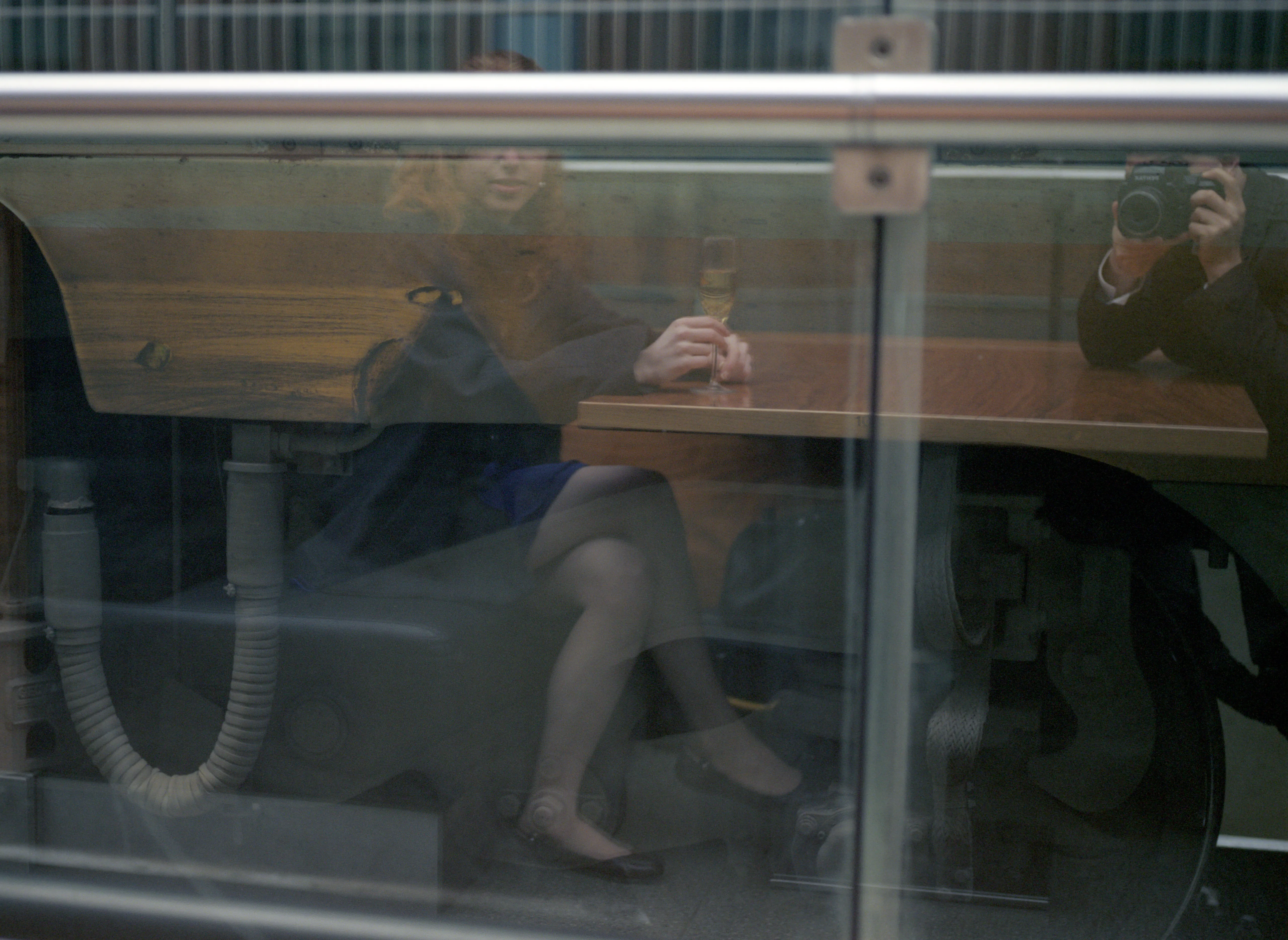 Clélia seen through a train window, seated at a carriage table, the photographer's reflection caught in the glass.