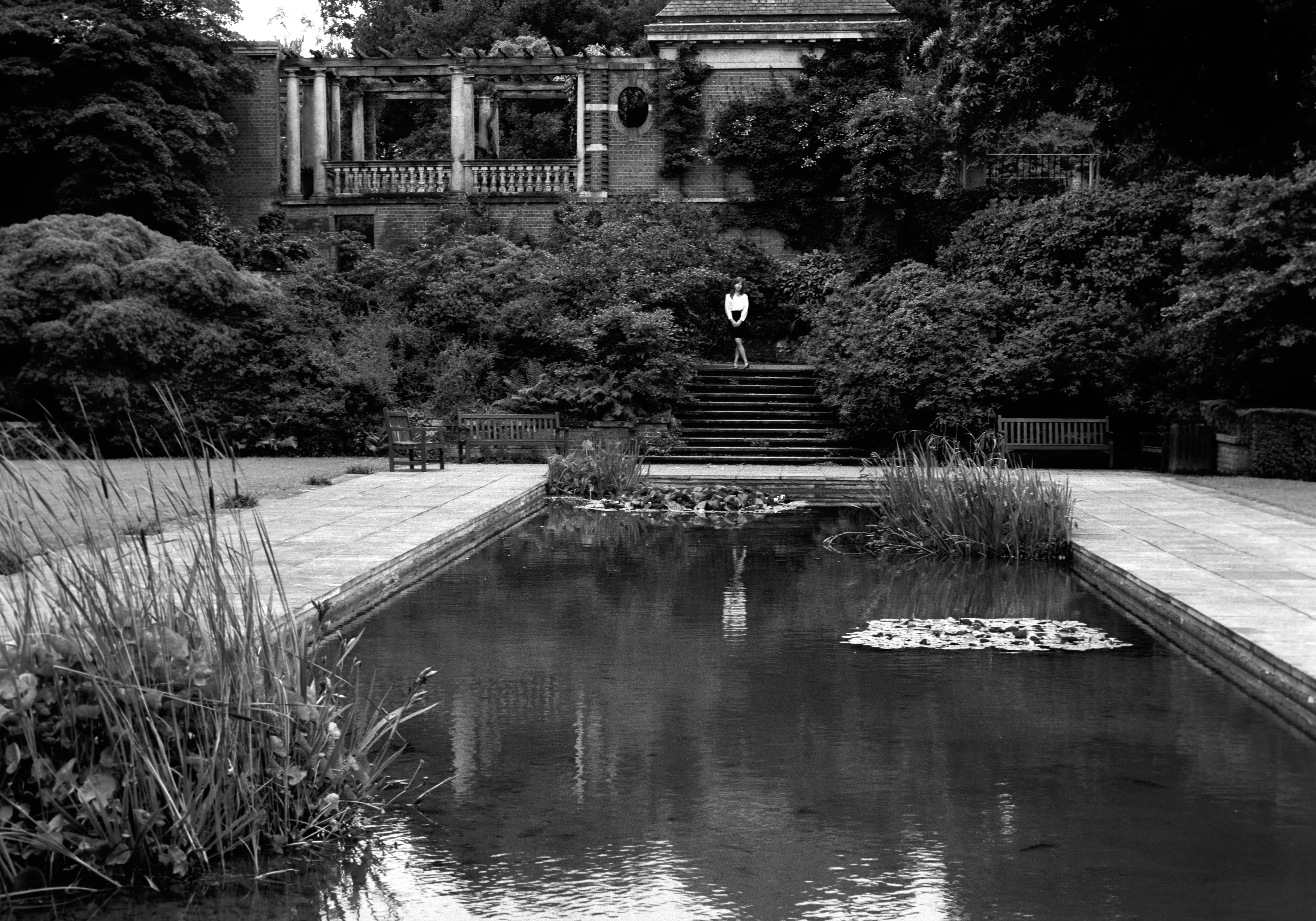 In black and white, a wide formal garden with a long reflecting pool and a colonnade beyond, Clélia a small figure in the distance.