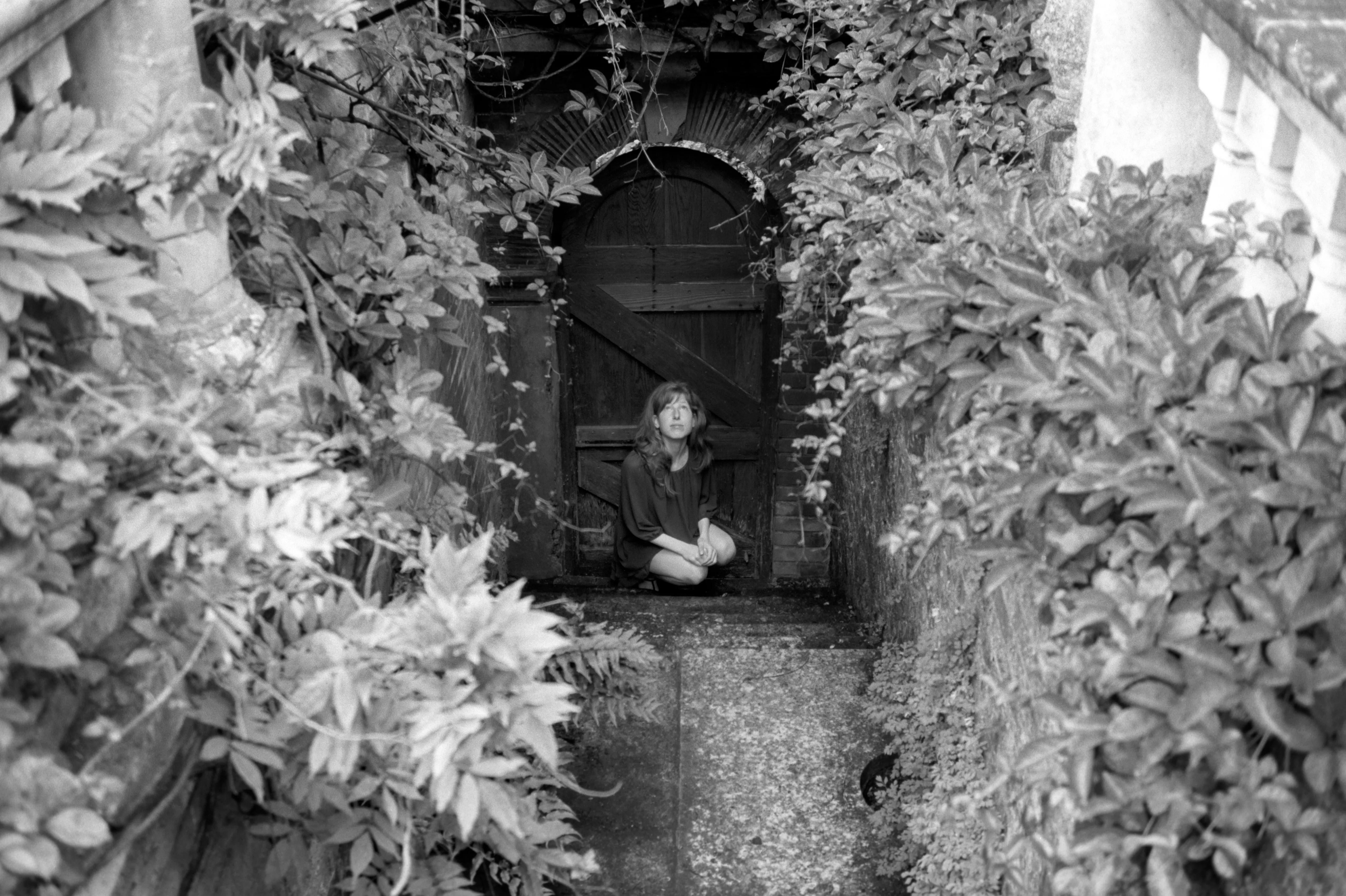 In black and white, Clélia sits small in an arched doorway framed by dense foliage, seen from a distance.