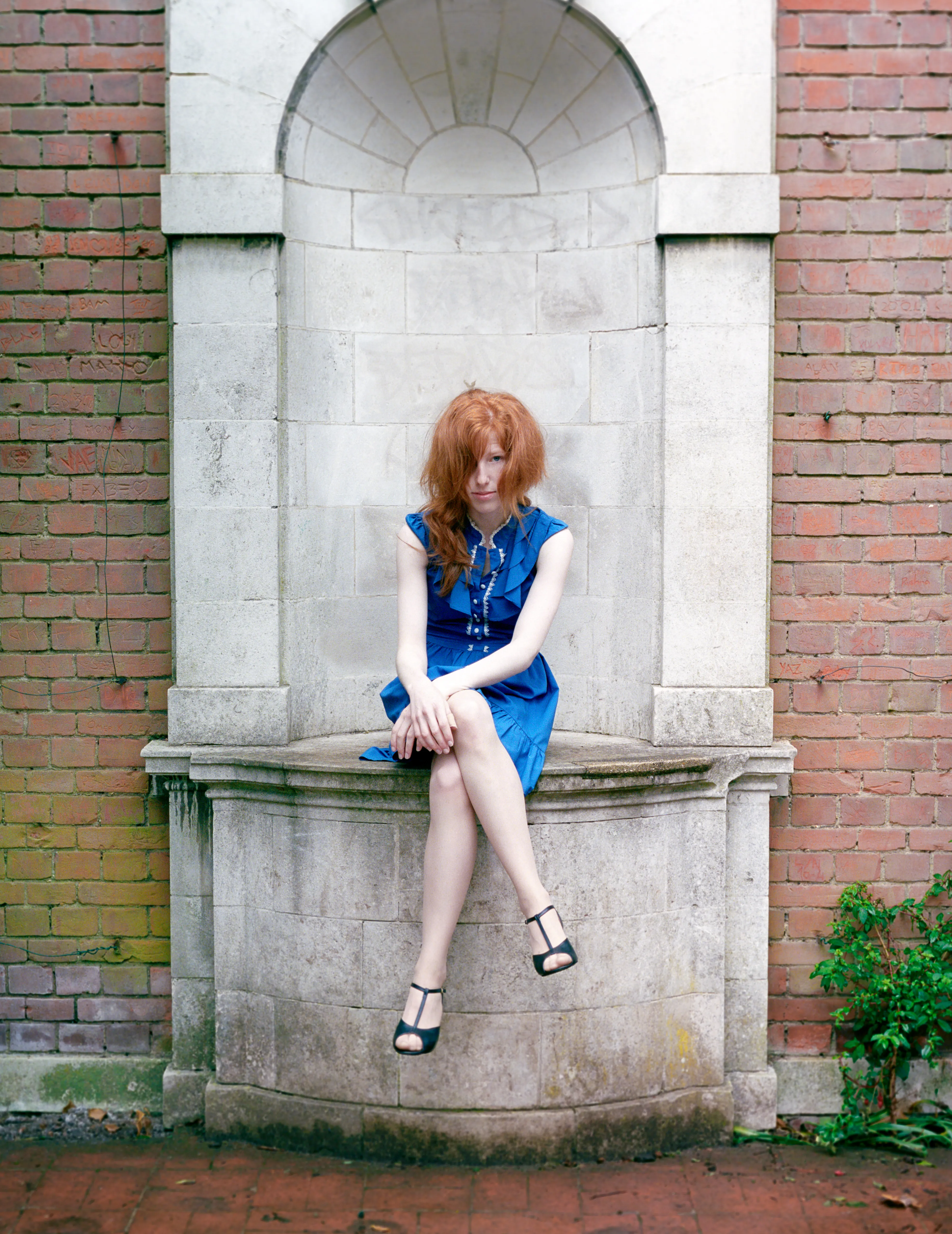Clélia sits in an arched stone niche set into a red-brick wall, in a blue dress.