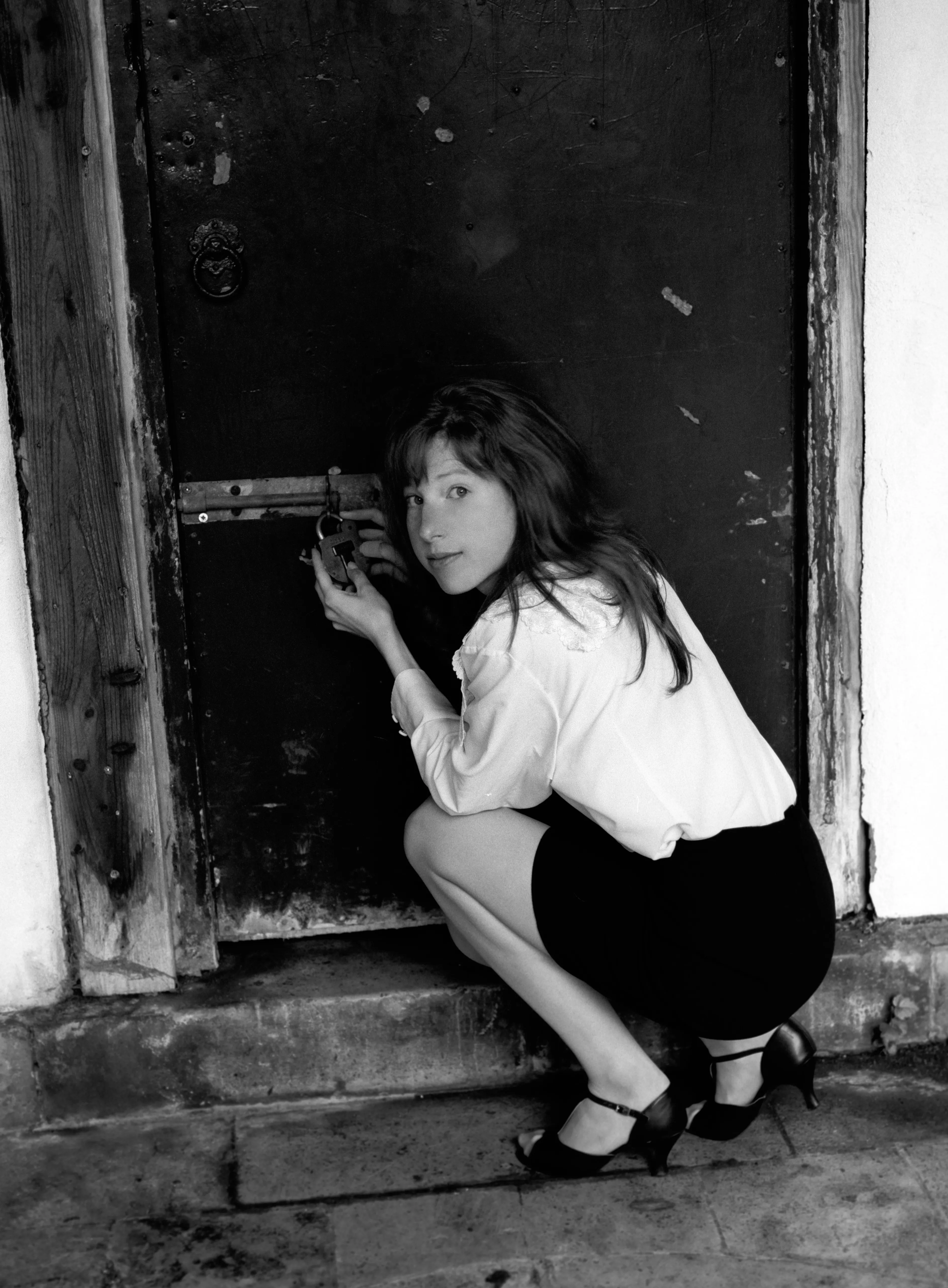 In black and white, Clélia crouches in the mouth of a weathered doorway, in a white shirt and dark skirt.